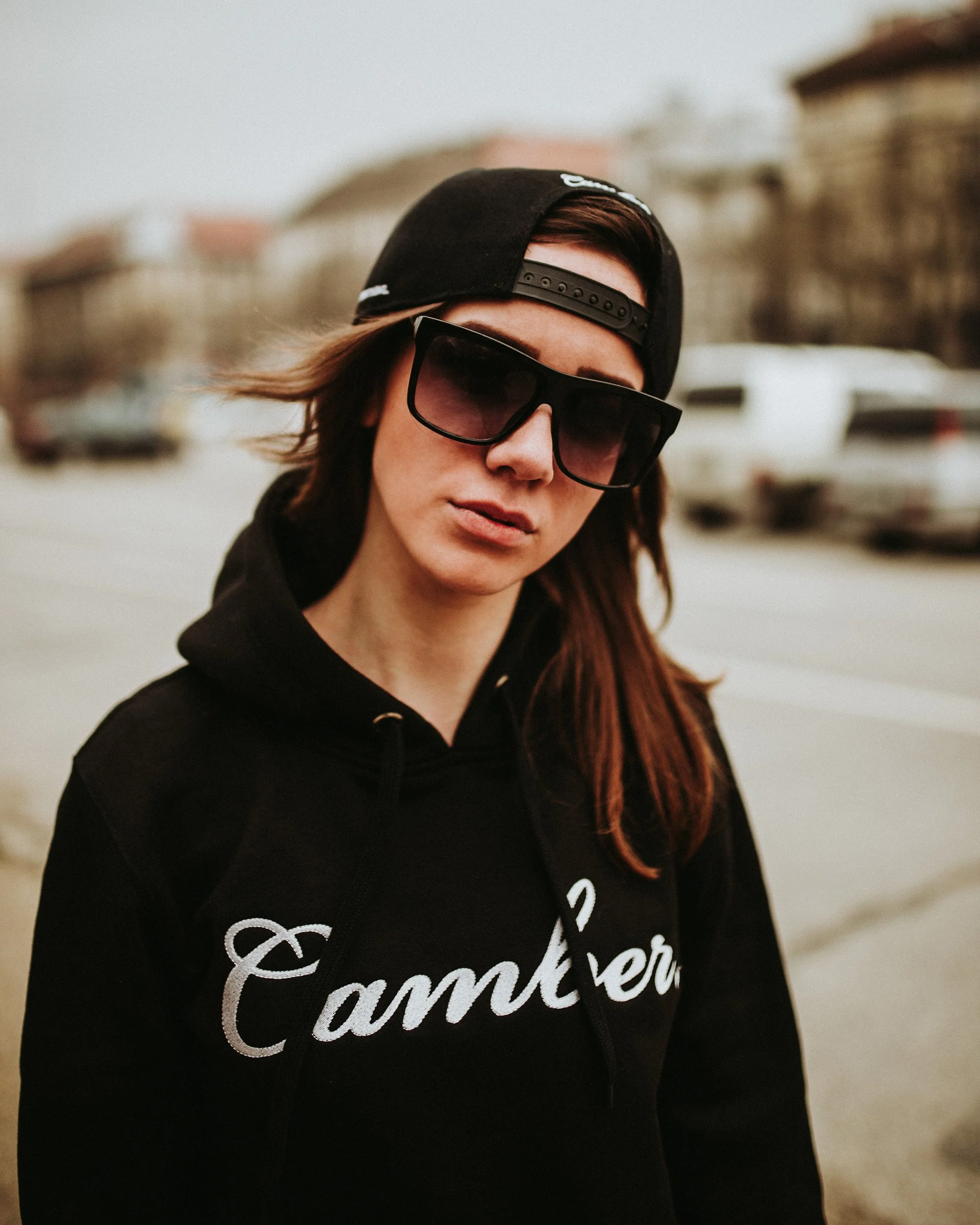 A woman with long reddish-brown hair wearing a black baseball cap backwards, dark sunglasses, and a black sweatshirt with white text, standing outdoors with blurred cars and buildings in the background.