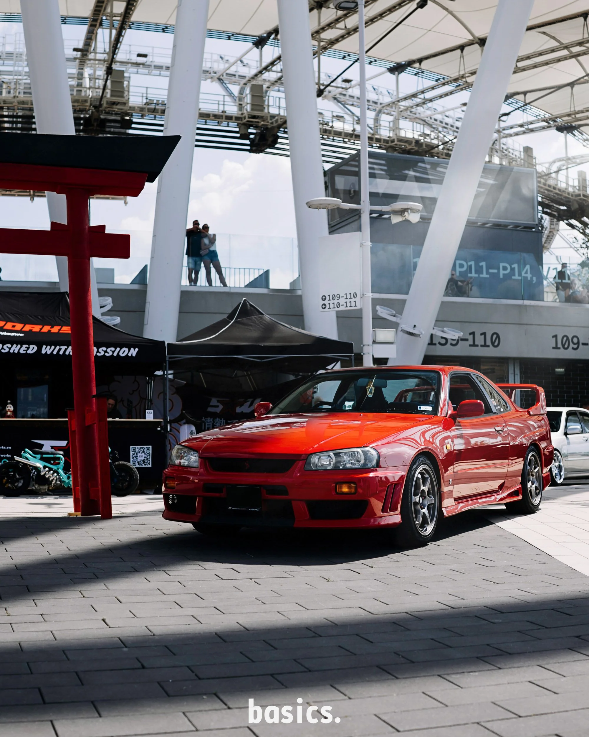 A bright red sports car with a sleek design parked on a paved area outside a modern structure with white support beams and a large glass viewing platform. Two people are seen on the platform in the background, with a sky filled with a few clouds abov