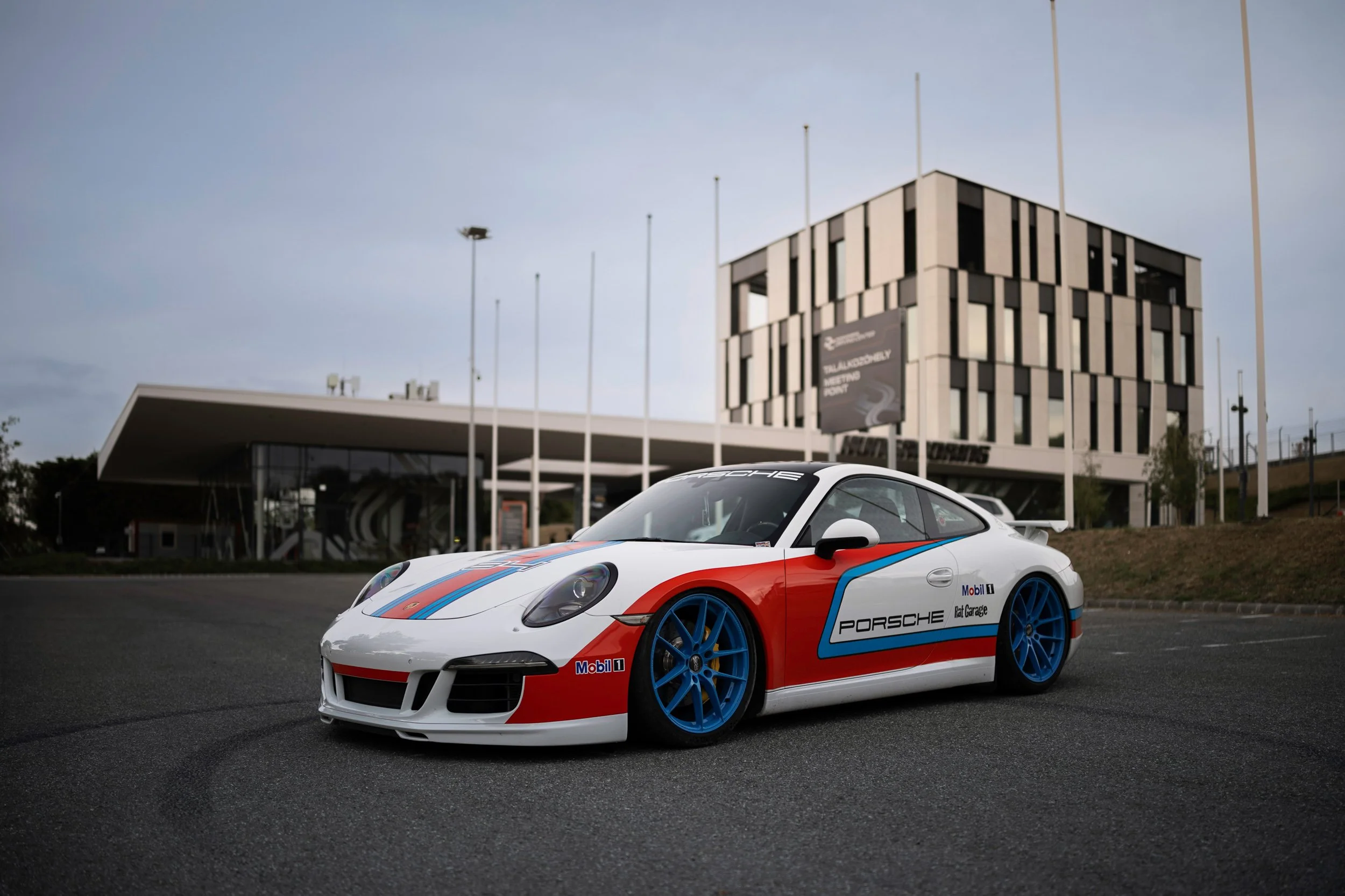 A white Porsche race car with blue and red racing stripes and branding, parked outdoors in front of a modern building with multiple flagpoles.