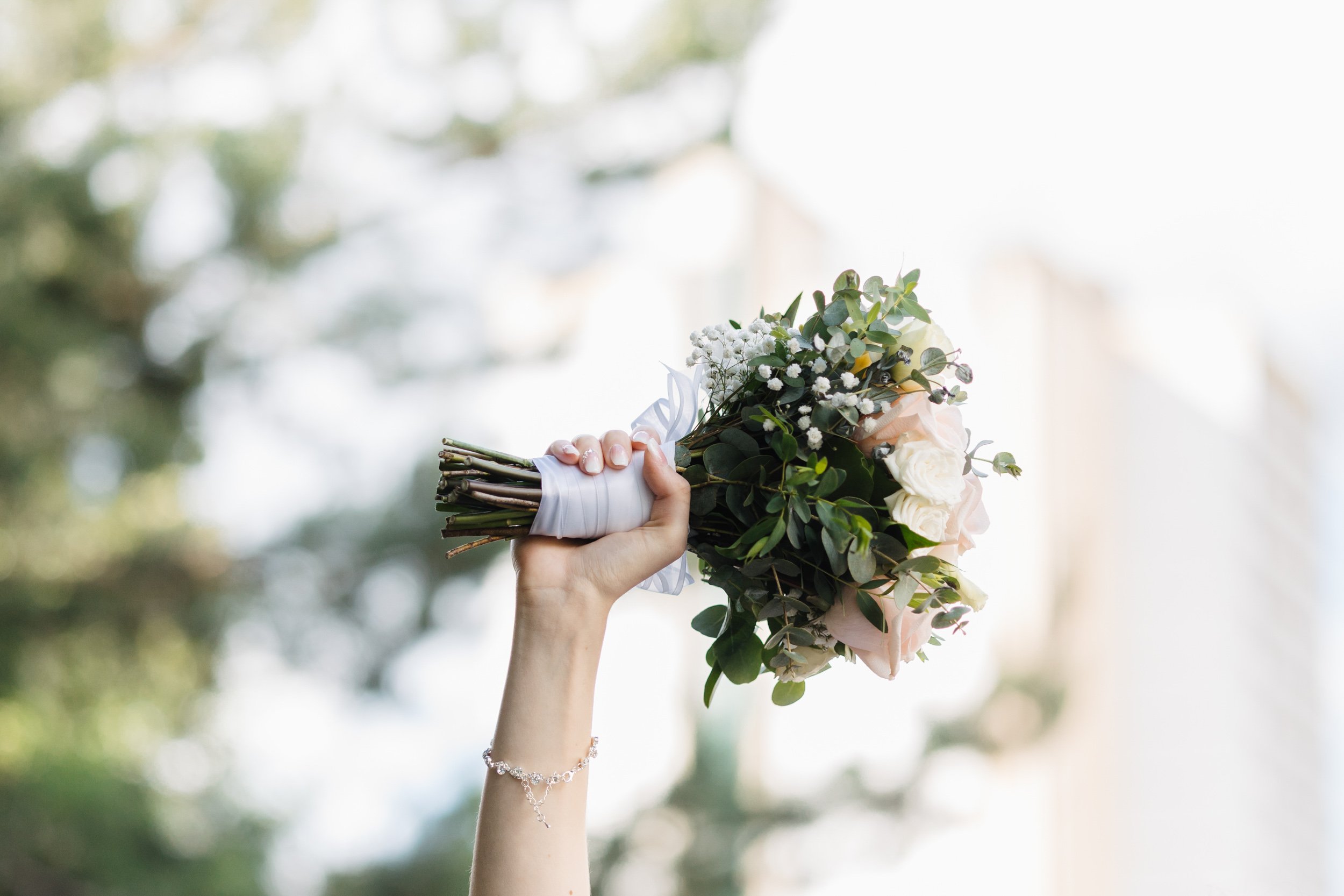 Hand holding a bridal bouquet of white roses, baby's breath, and green foliage against a blurred outdoor background.