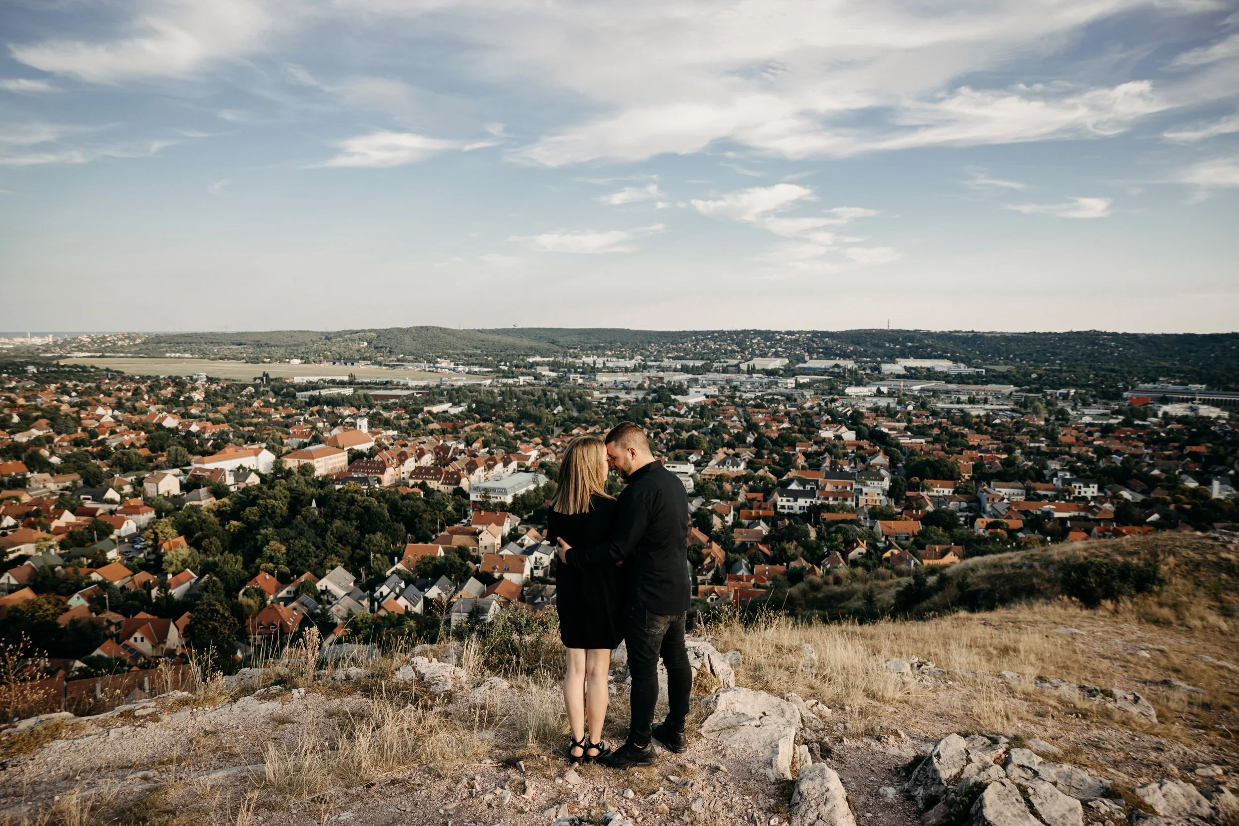 A couple standing close together on a grassy hill with rocks, overlooking a city with many houses and industrial buildings, under a partly cloudy sky.