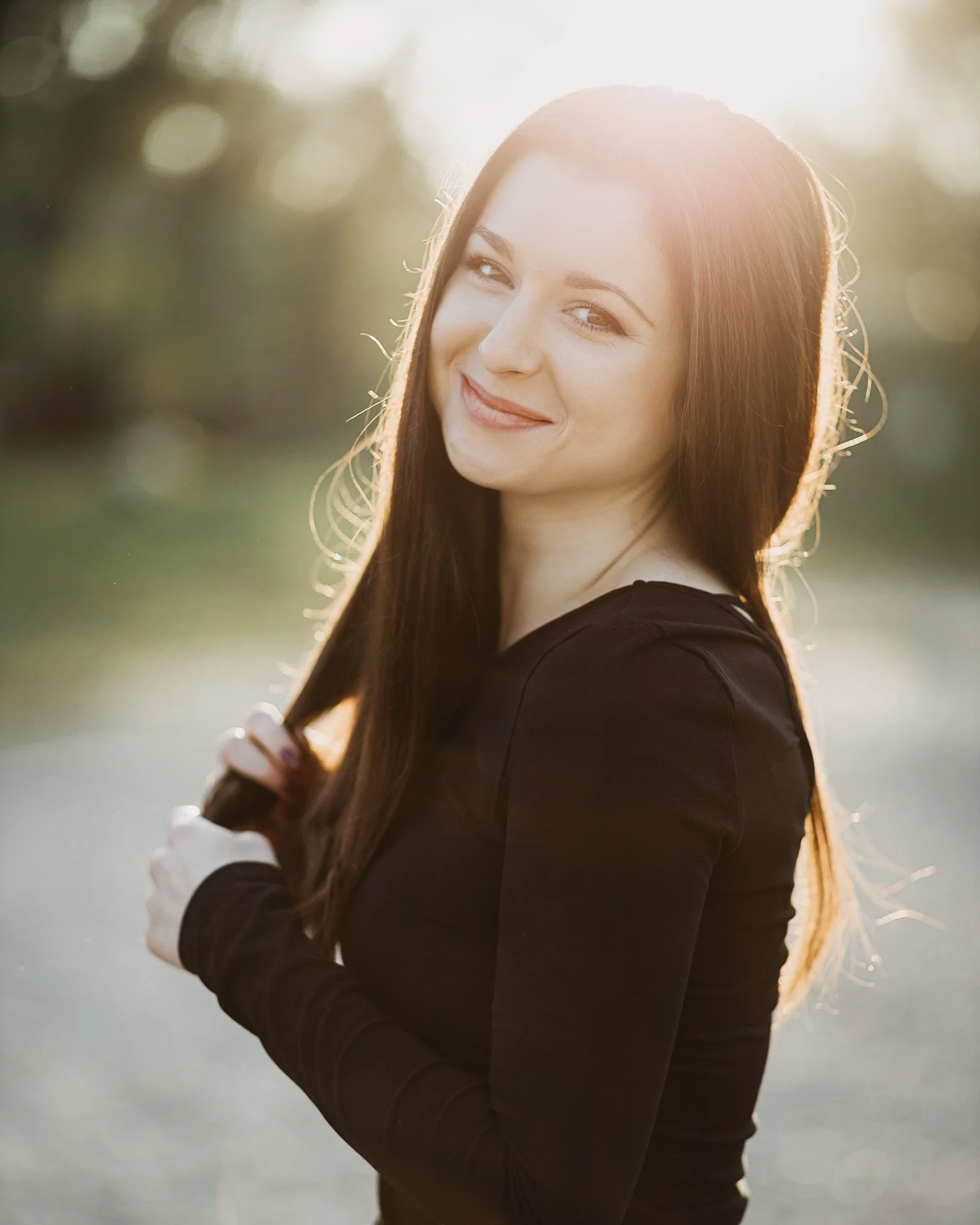 A woman with long brown hair smiling outdoors in sunlight, wearing a black top.