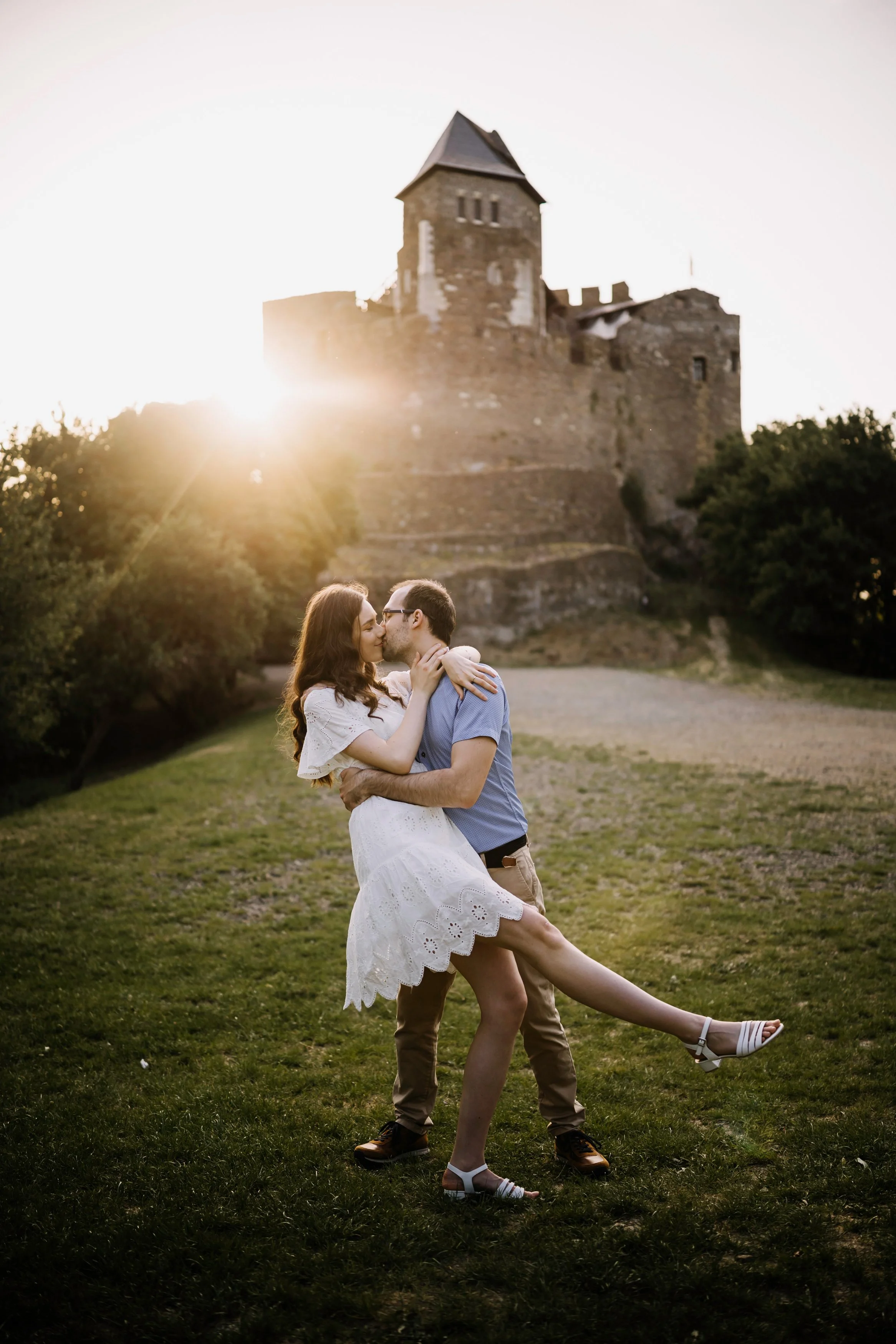 A young couple sharing a kiss outdoors at sunset, with a castle on a hill in the background.