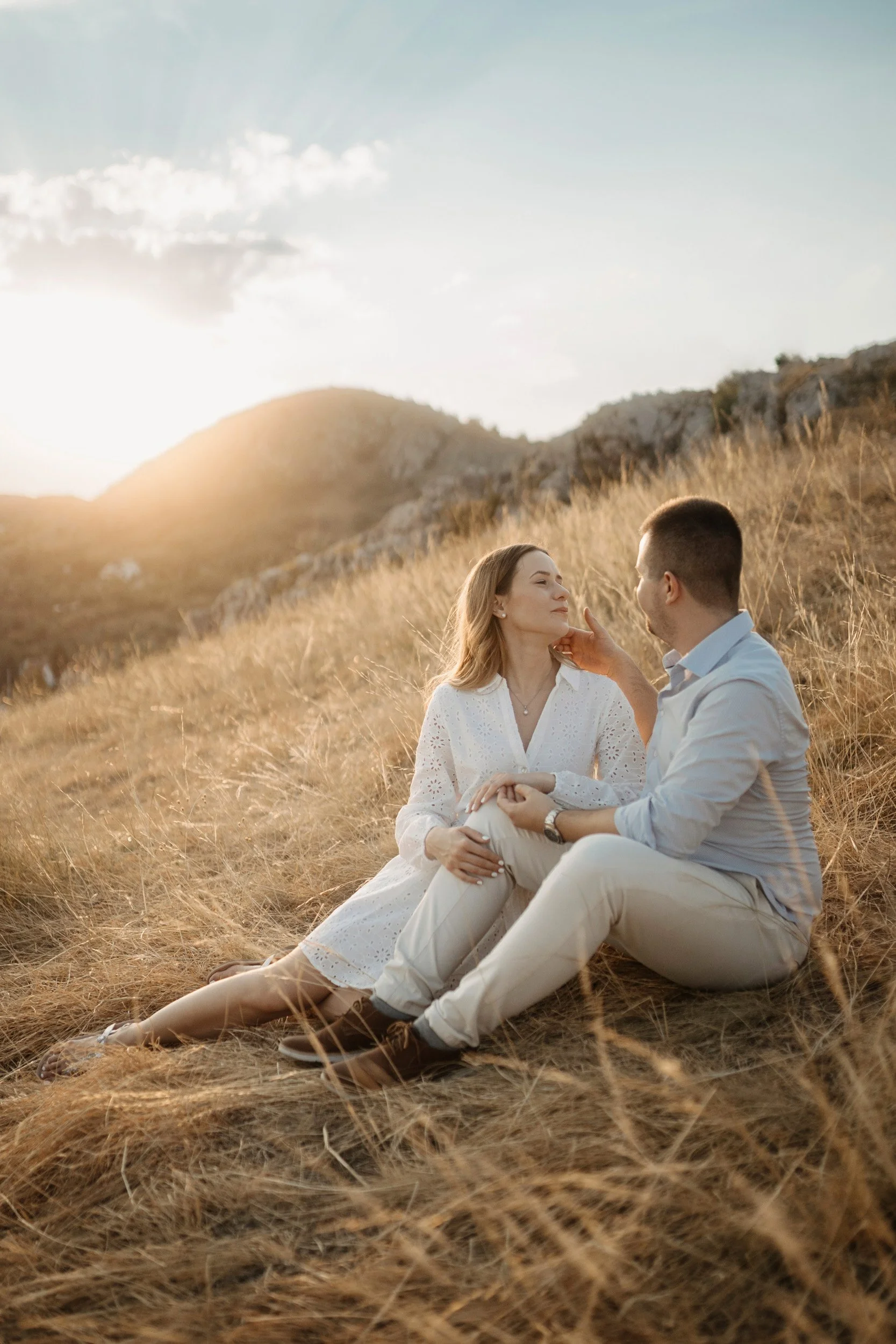 A couple sitting in a field of tall, dry grass with mountains in the background at sunset. The man is touching the woman's chin, and they are looking into each other's eyes.