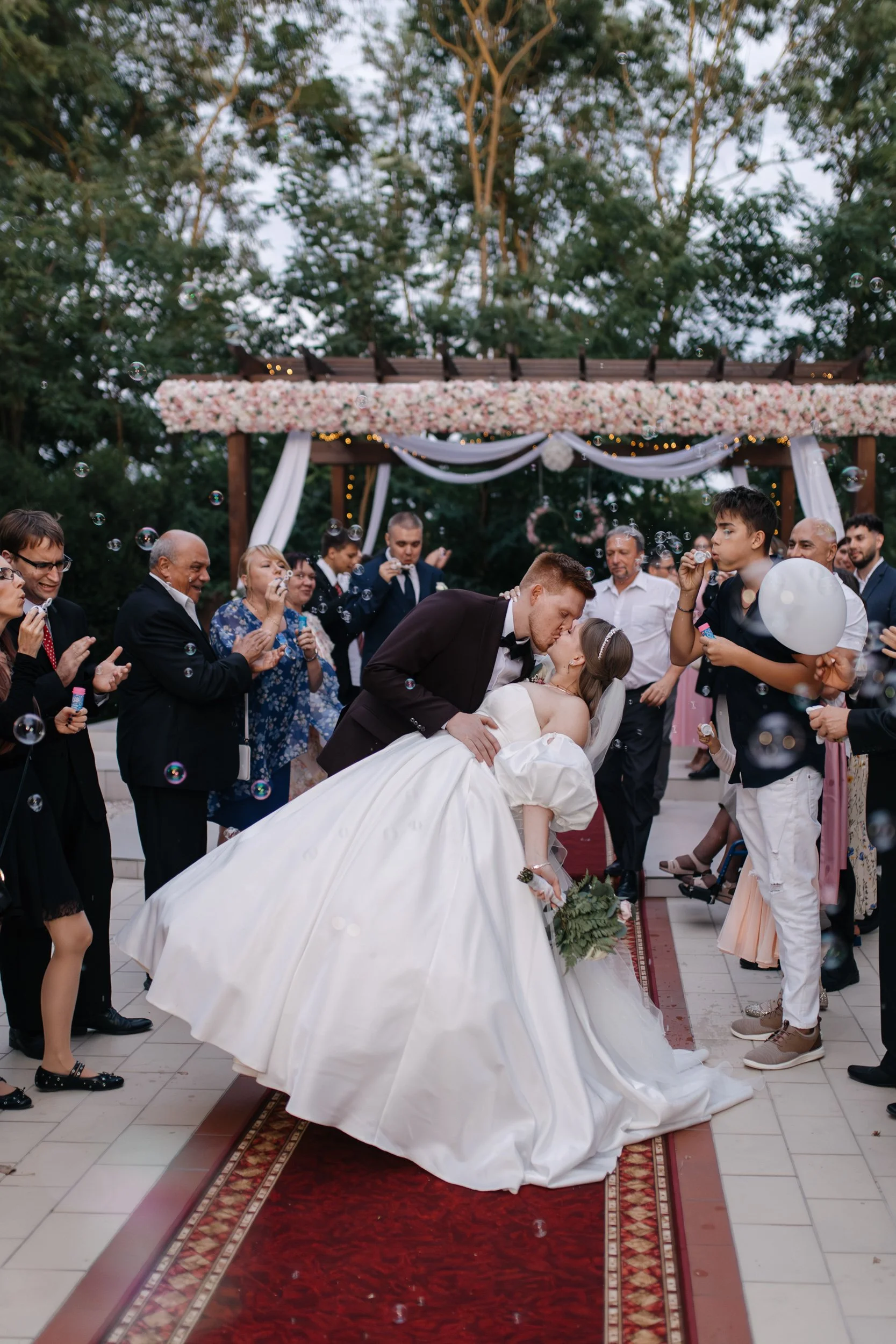 A wedding celebration with a bride and groom dancing, surrounded by guests outdoors under a decorated archway with flowers and string lights.