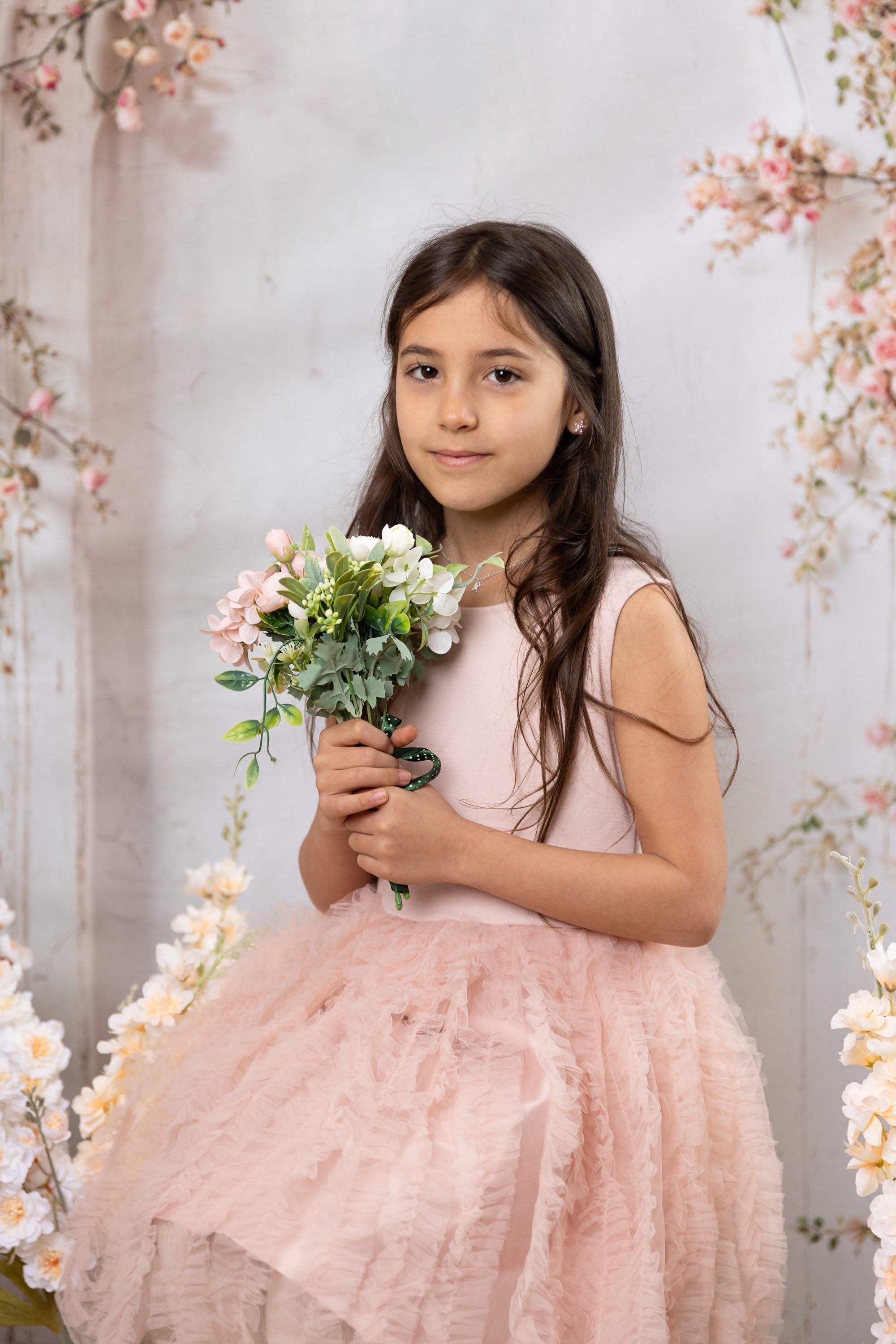 Young girl in pink dress holding a bouquet of flowers, standing in front of a floral backdrop.