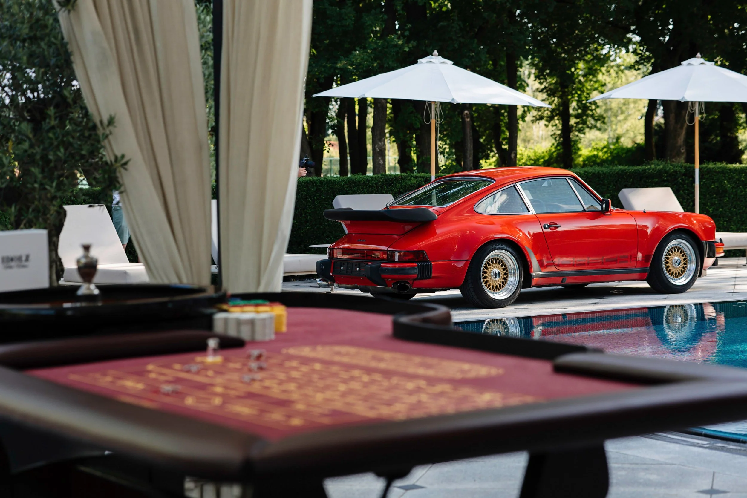 A red classic Porsche 911 parked outdoors under white patio umbrellas, with trees in the background. The photo is taken from inside a building near a gaming or poker table.