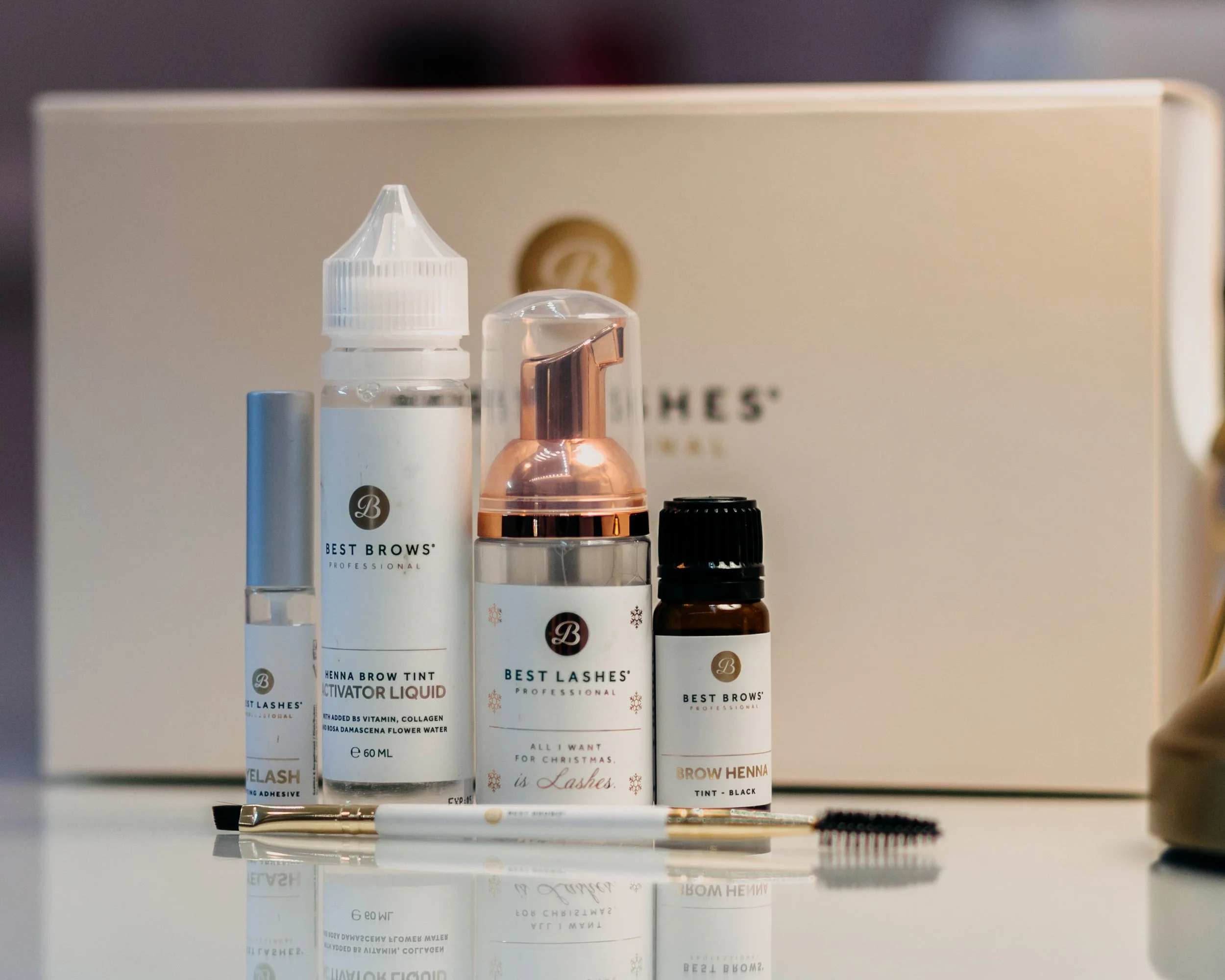 Various beauty and skincare products on a reflective surface, including sprays, bottles, and a small tube, with a beige background and some branding visible.
