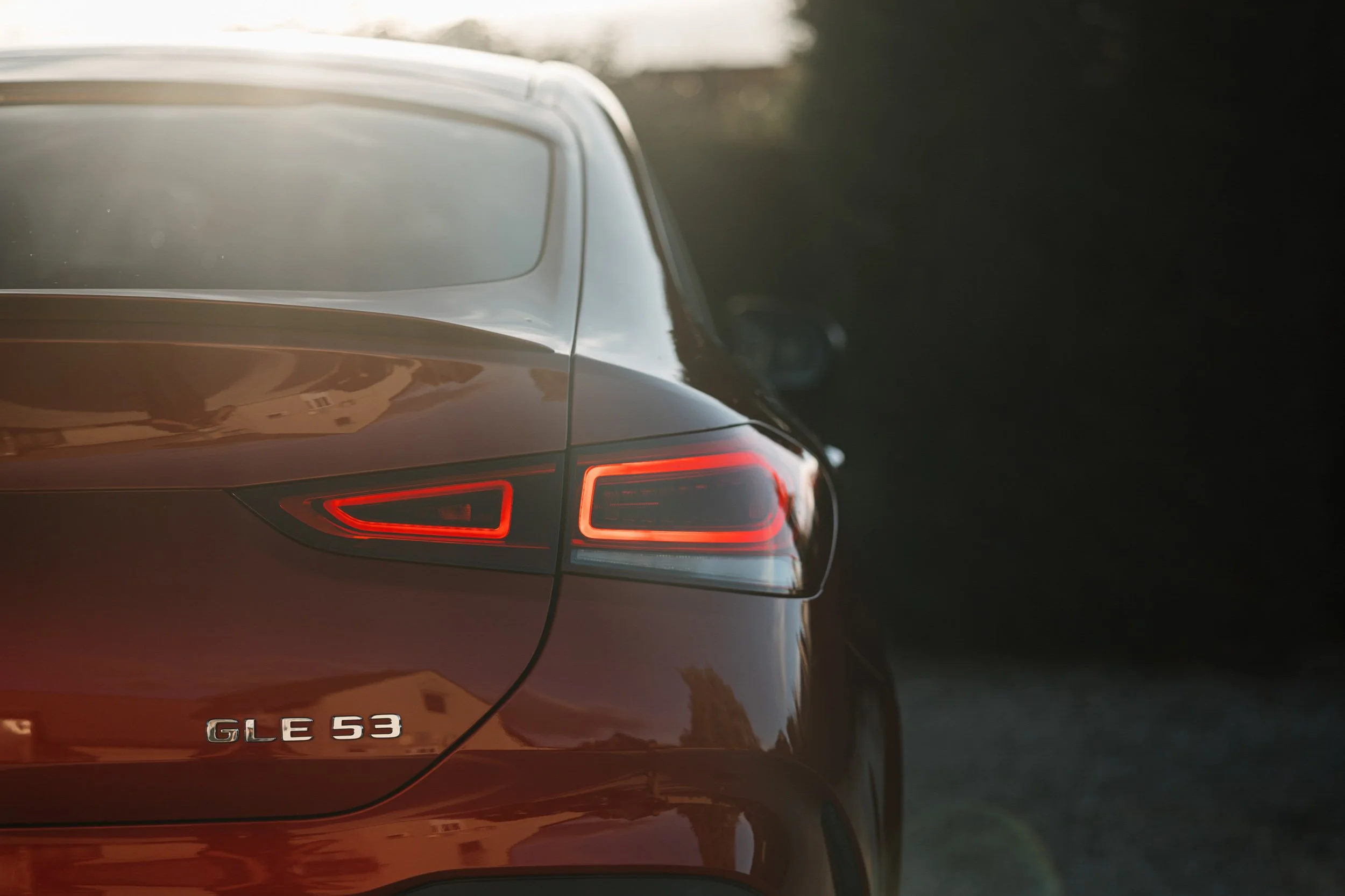 The rear of a gray luxury car with taillights illuminated, displaying the badge "GLE 53" on the trunk, parked outdoors with dark surroundings.