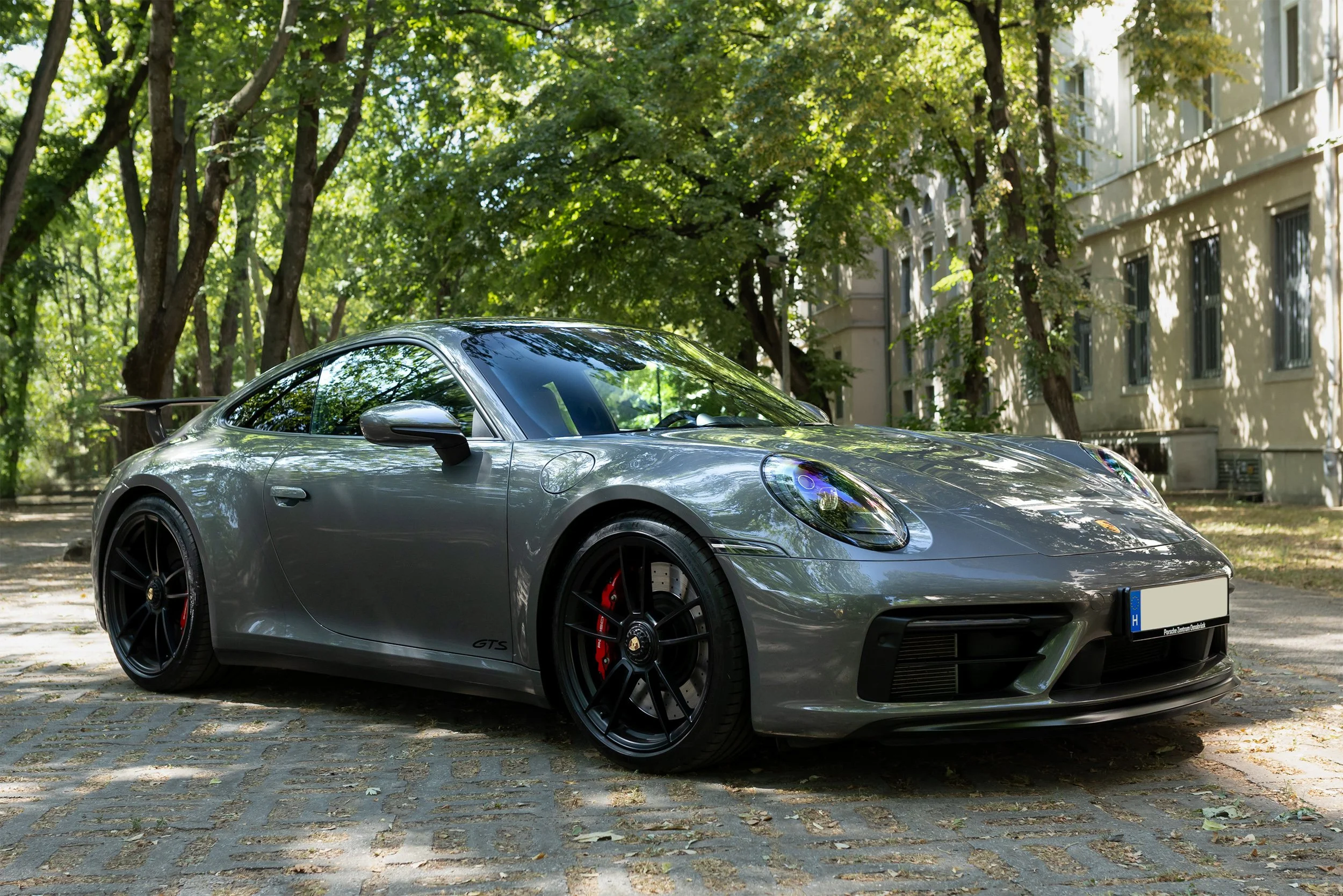 A silver Porsche sports car parked on a stone-paved street under trees with green foliage, and apartment buildings in the background.