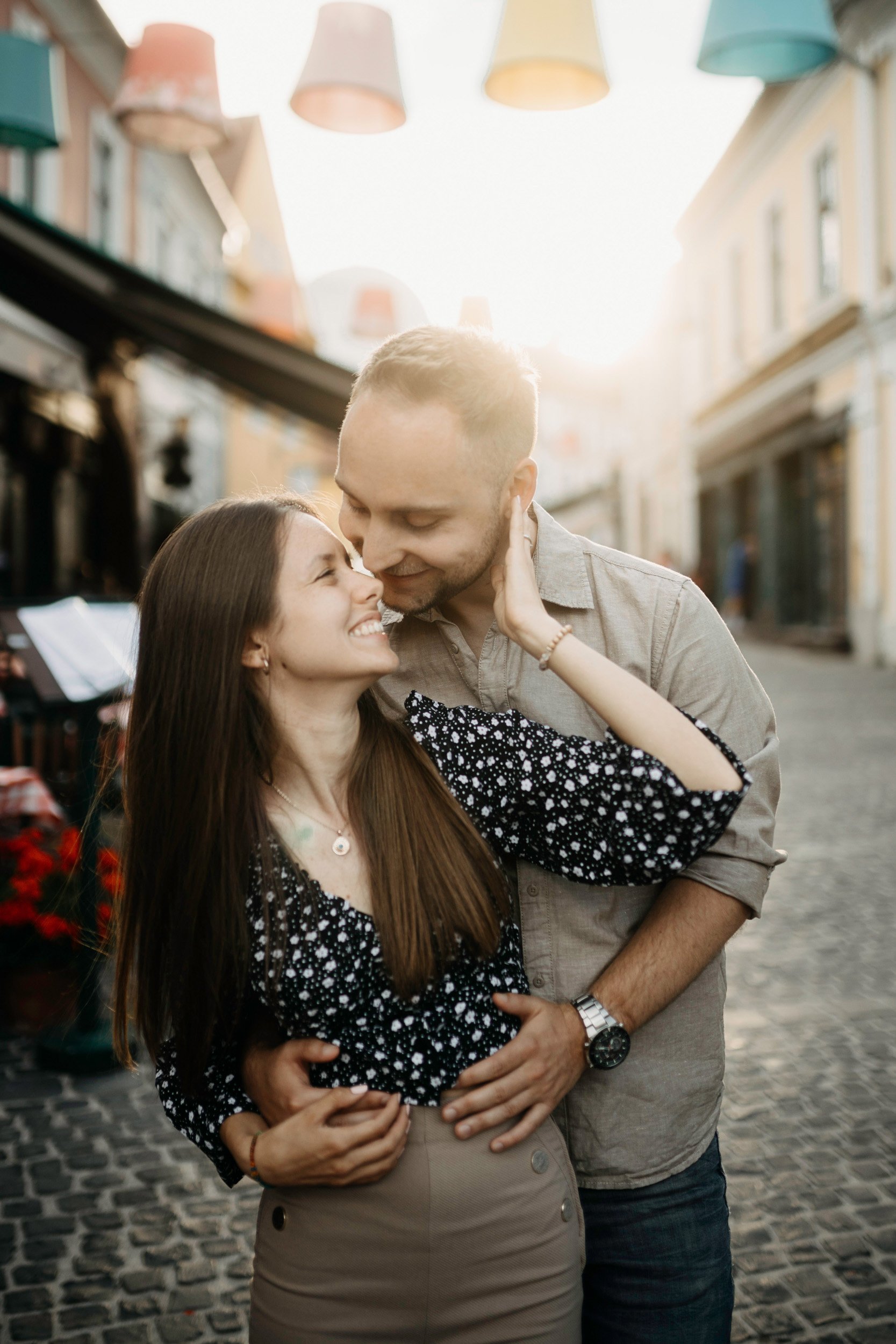A couple embracing and smiling in a street with colorful buildings and hanging lamps, during the daytime.