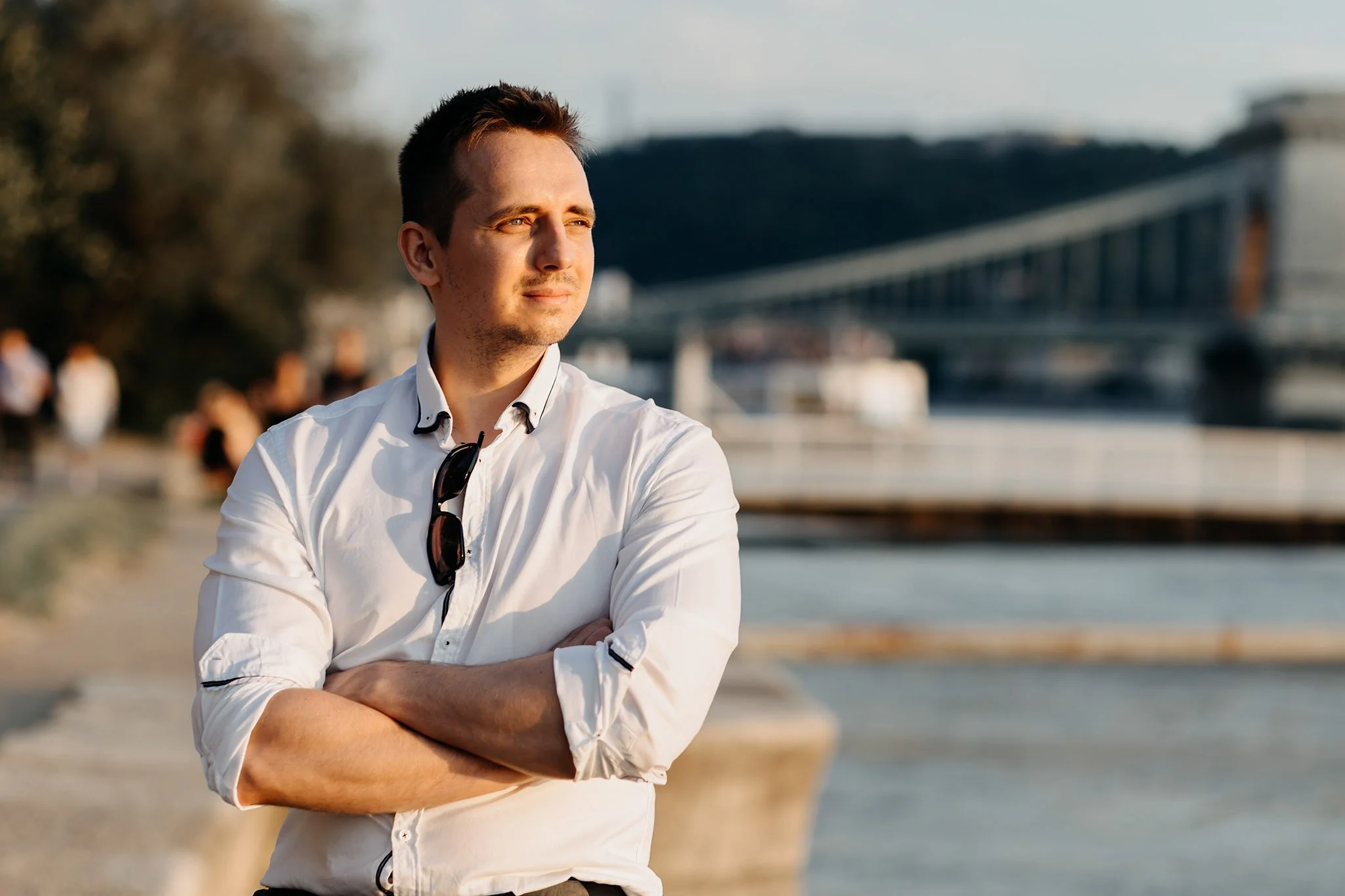 A young man with short dark hair standing outdoors by a waterfront, wearing a white shirt with sunglasses hanging from the collar, crossing his arms, and looking into the distance.