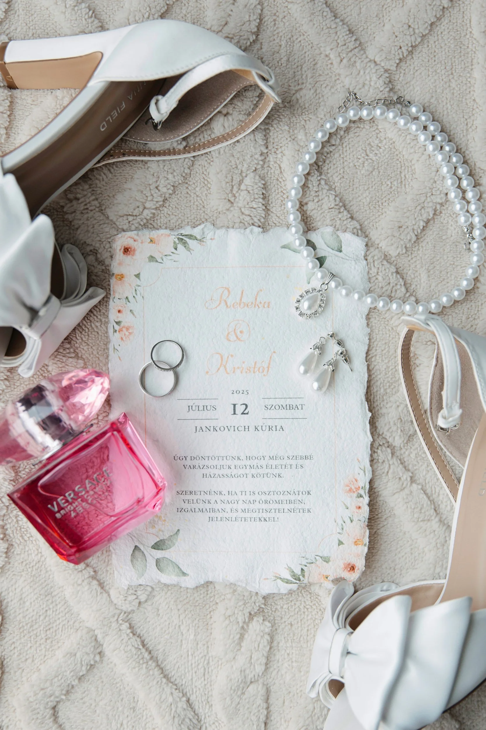 Wedding invitation, pearl necklace with pendant, rings, pink perfume, white shoes with ribbon, beige purse on a soft beige textured blanket.