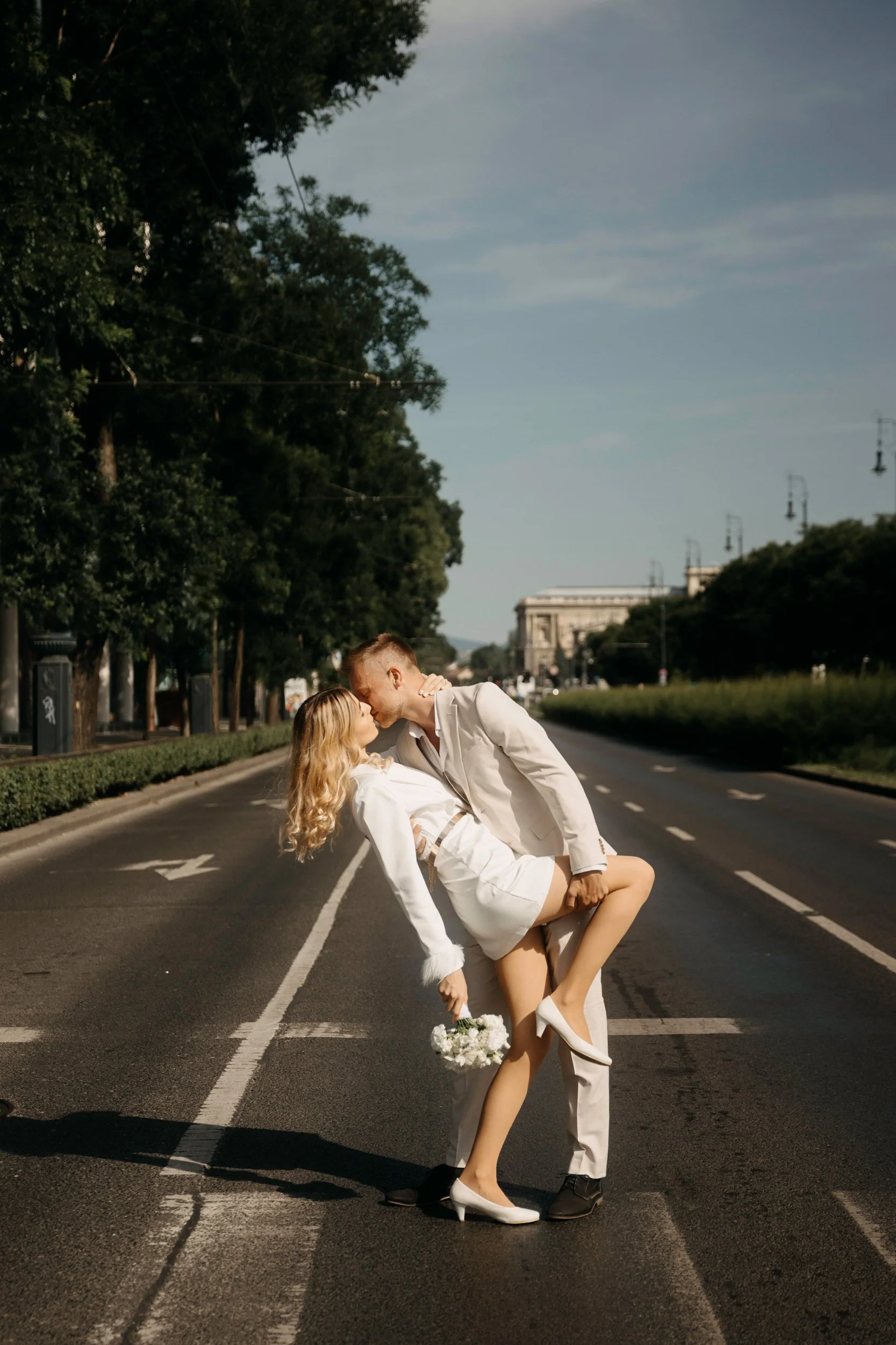 A couple in white clothes sharing a romantic moment on an empty city street, with the man dipping the woman and holding her close, the woman holding a small bouquet of white flowers.
