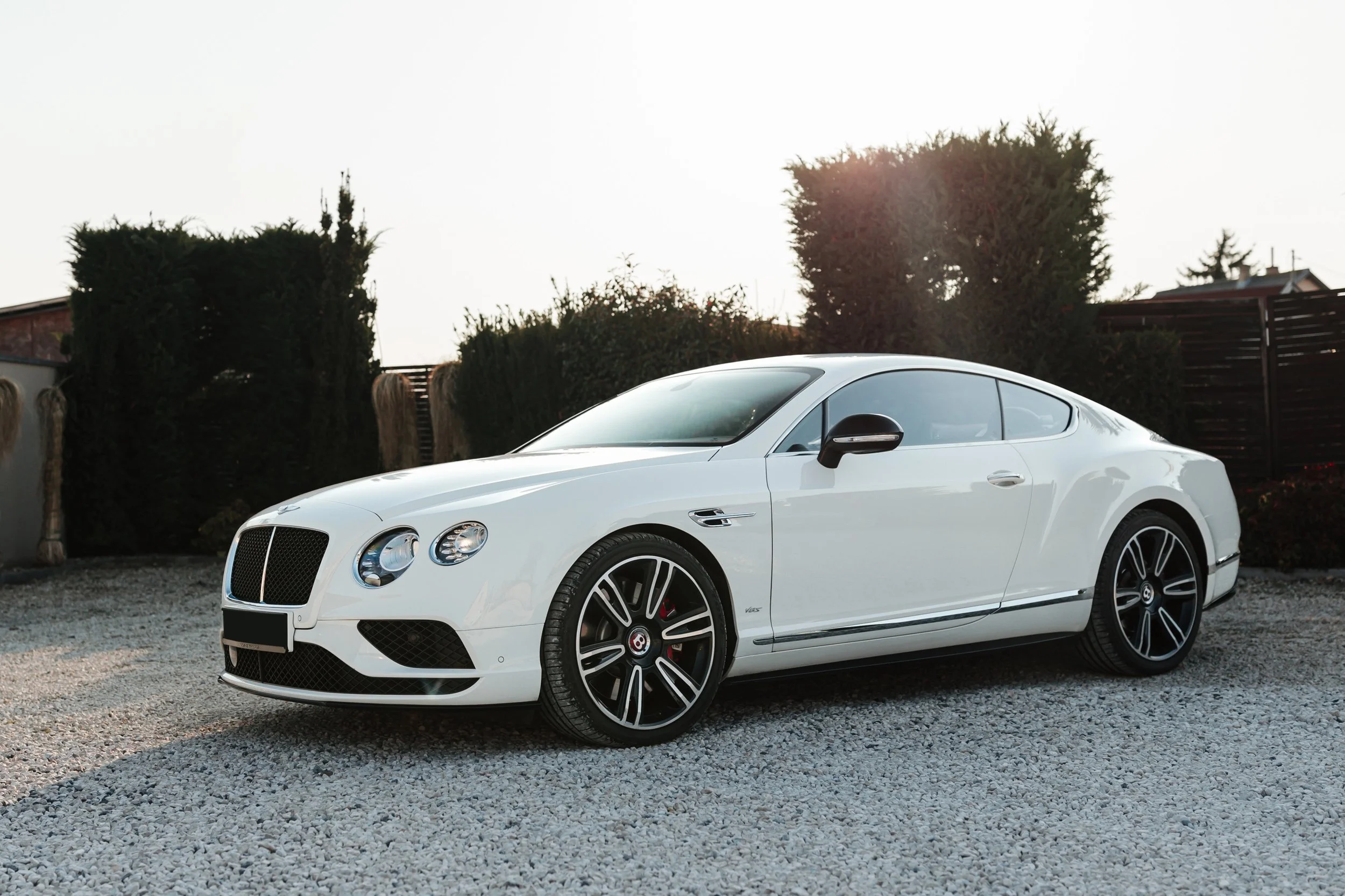 A white luxury coupe car parked on a gravel surface with green bushes and wooden fencing in the background during daylight.