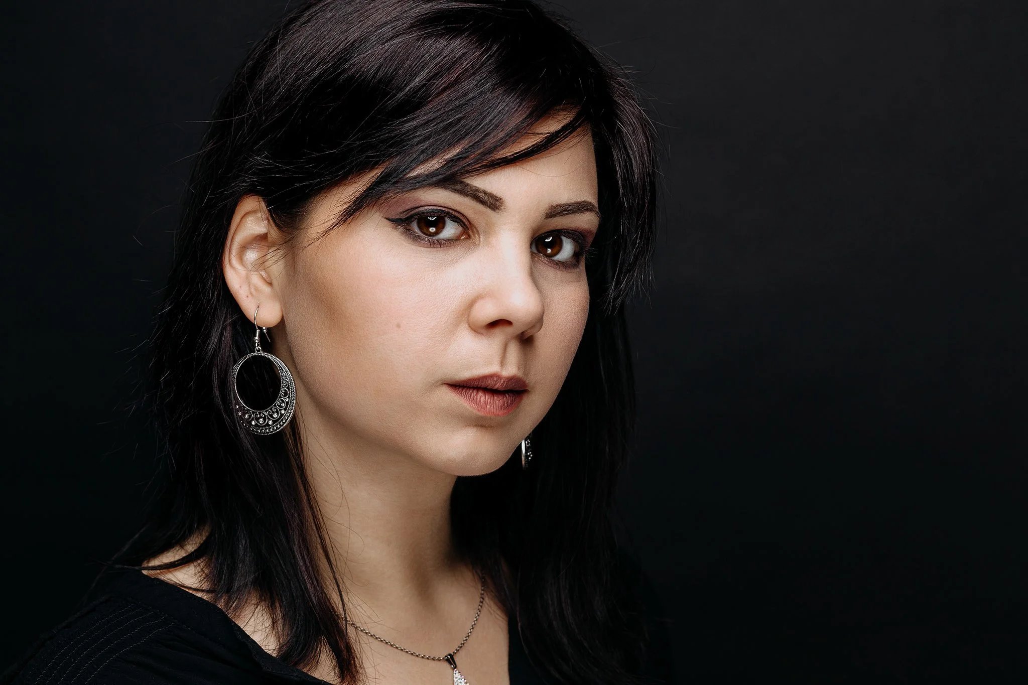 A woman with dark hair and brown eyes wearing earrings and a necklace, against a black background.