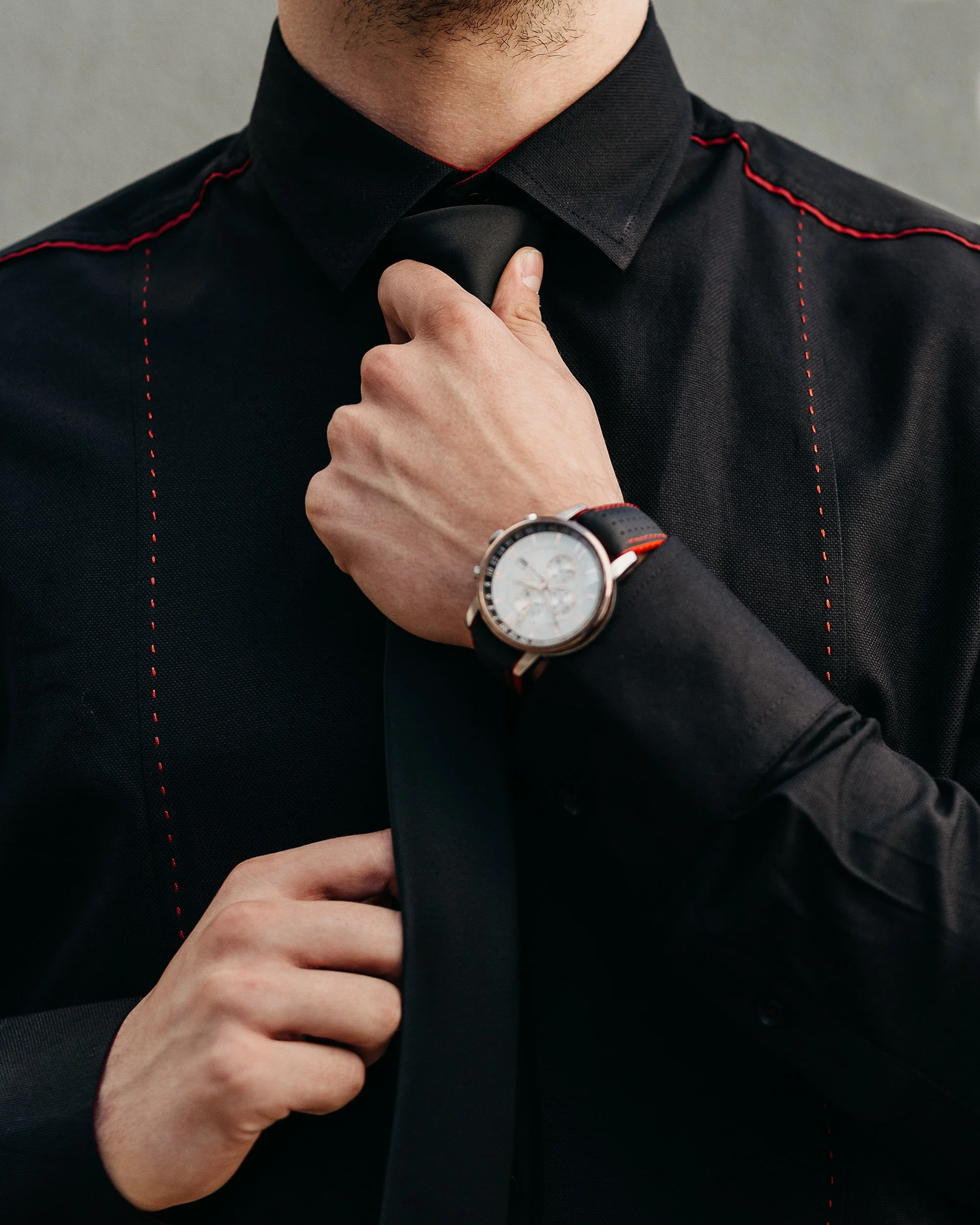 Close-up of a man adjusting his black necktie, wearing a black shirt with red stitching details, and a wristwatch with a white face and black leather strap.