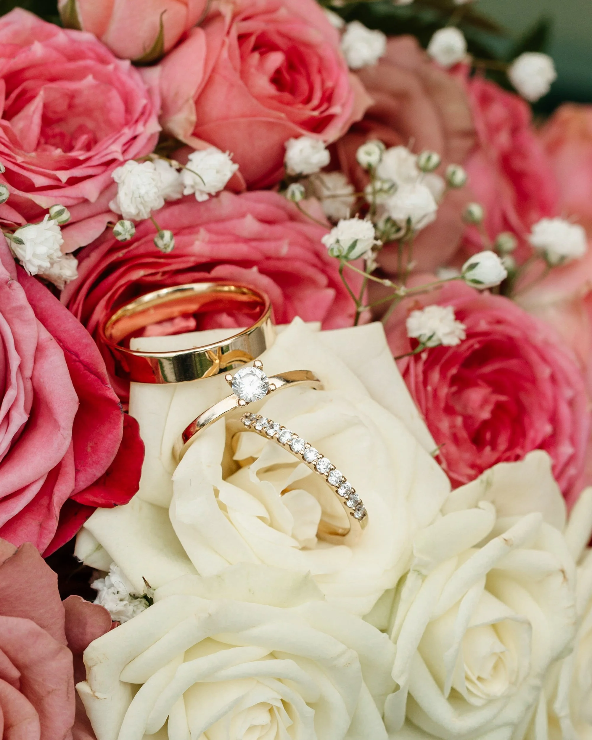Close-up of a bouquet with pink and white roses, small white filler flowers, and gold and silver wedding rings resting on white rose petals.