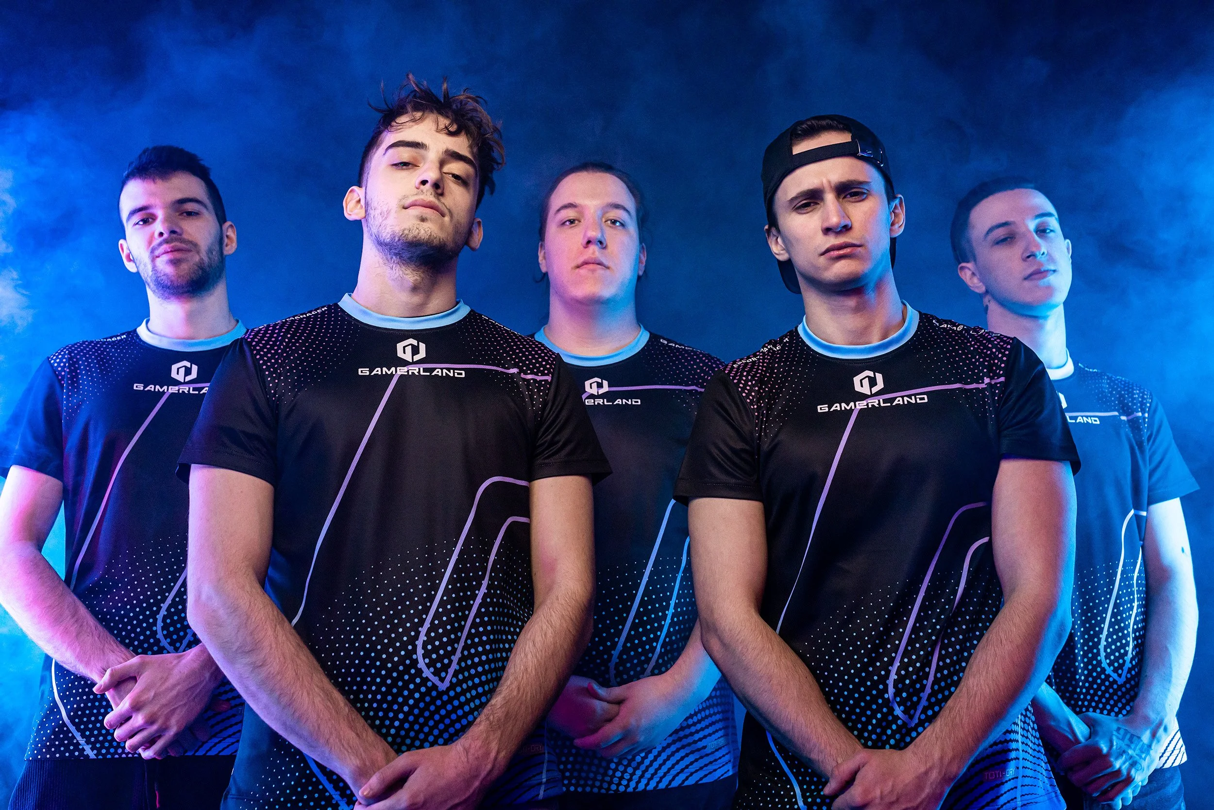 Group of five esports team members wearing black gaming jerseys with 'GAMERLAND' logo, standing against a blue smoky background.