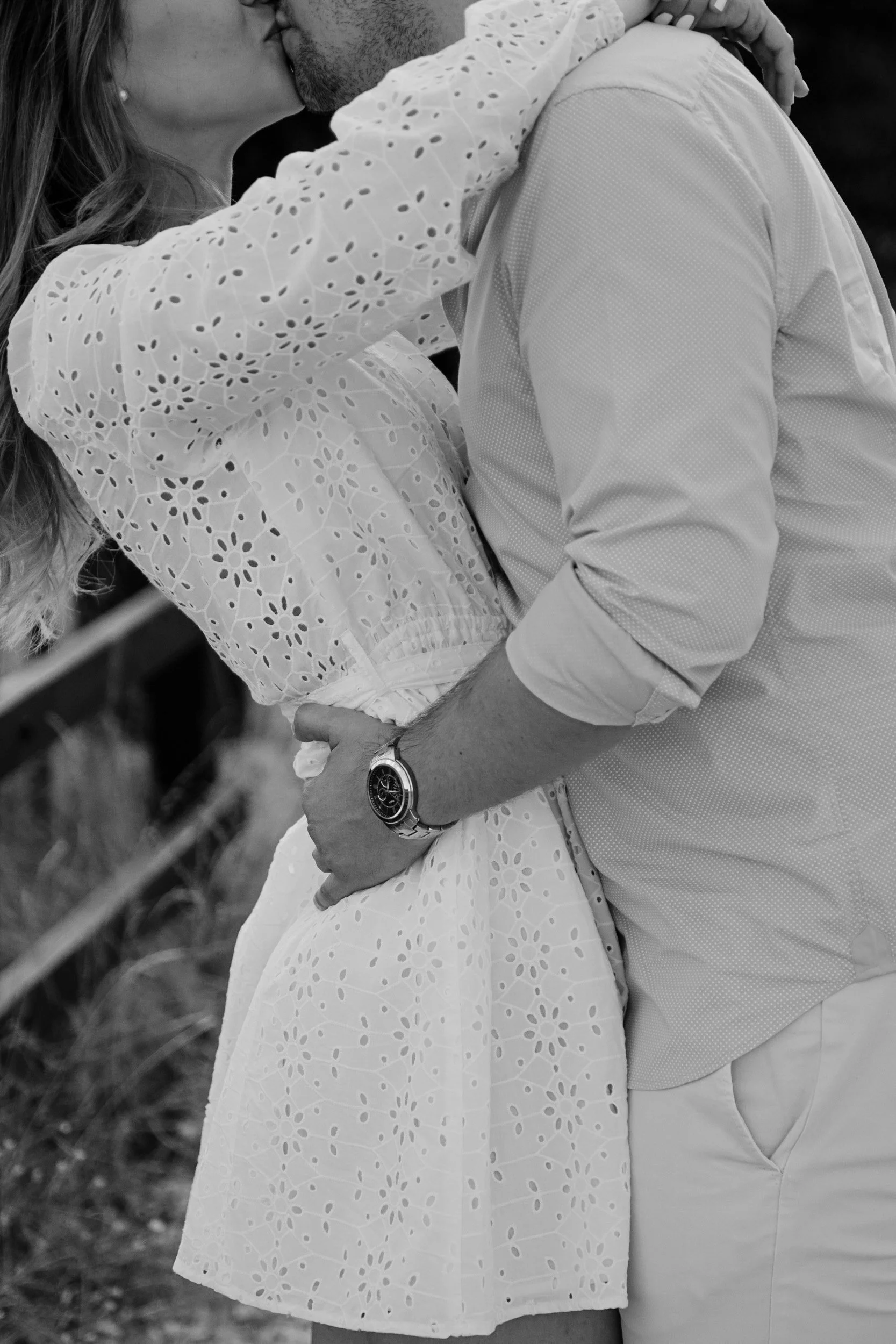 A couple is sharing a kiss outdoors, with the woman wearing a white eyelet dress and the man in a light-colored shirt and watch.