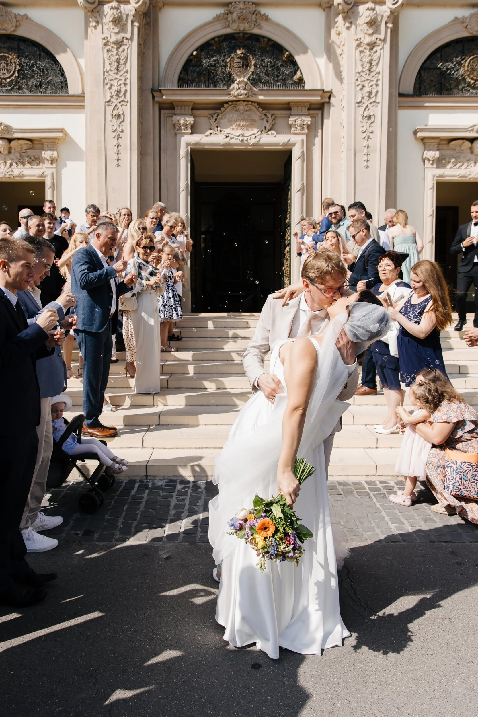 A bride and groom share a kiss on the steps of a grand, ornate building while surrounded by friends and family celebrating. The bride holds a colorful bouquet, and the groom wears glasses and a white suit.