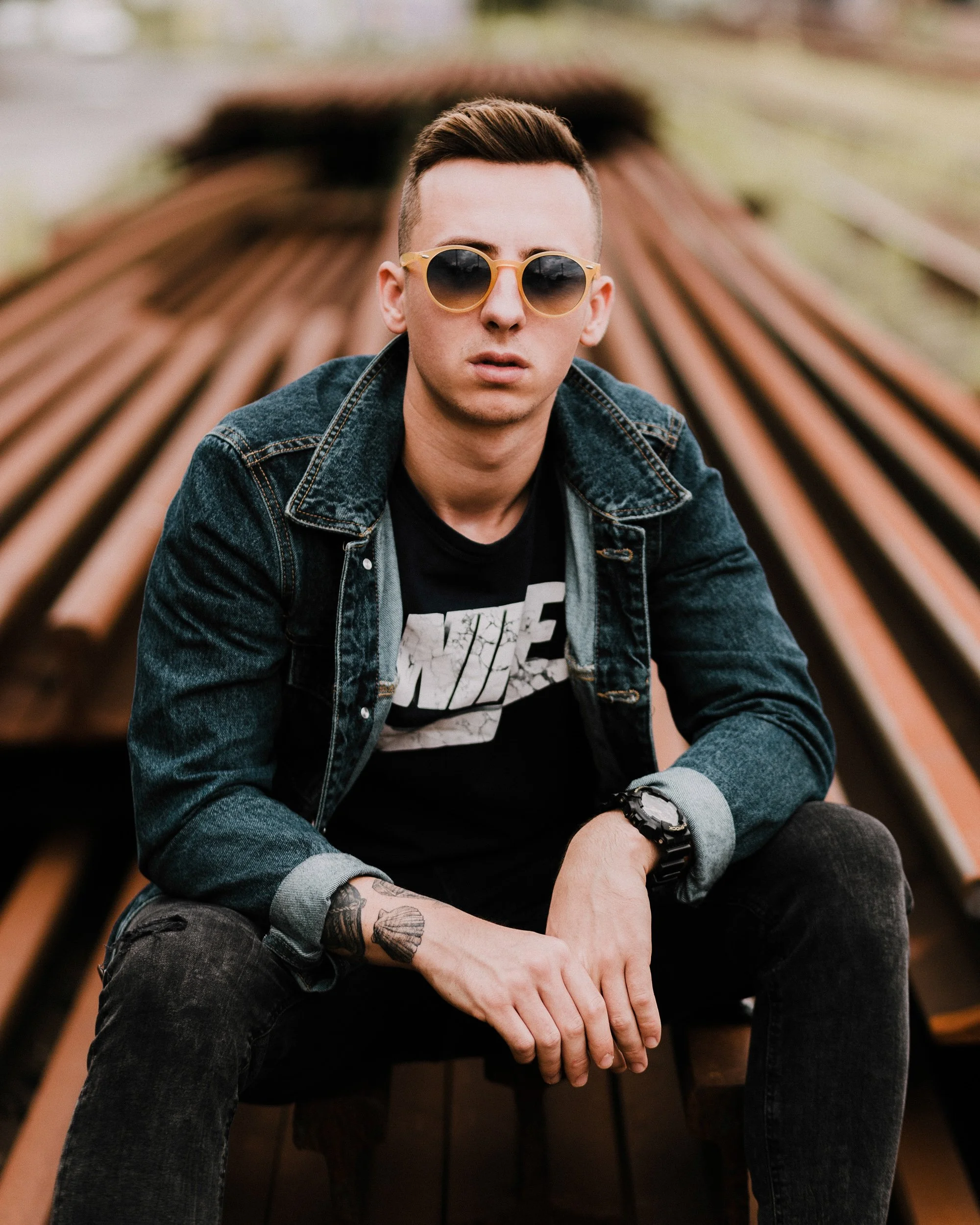 A young man with short brown hair, wearing sunglasses, a denim jacket over a black graphic t-shirt, and a black watch, sitting on what appears to be a railroad track with blurred wooden ties in the background.