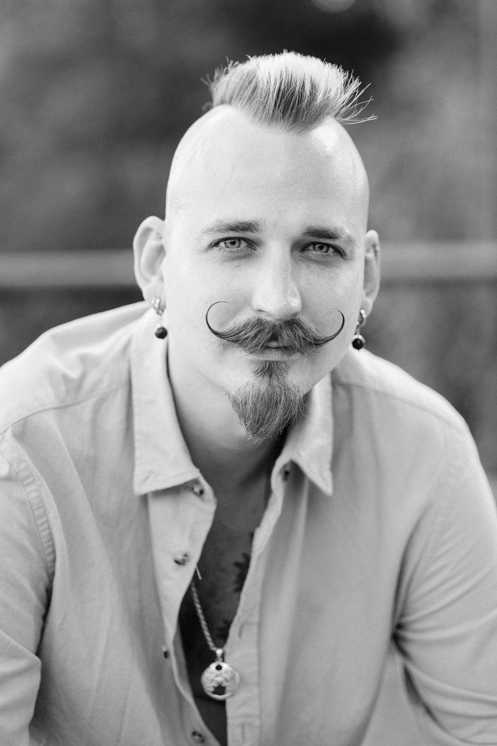 Black and white photo of a man with a distinctive hairstyle, styled in a high pompadour with shaved sides, facial hair including a styled mustache and goatee, earrings, and a necklace with a skull pendant, wearing a partially unbuttoned shirt.
