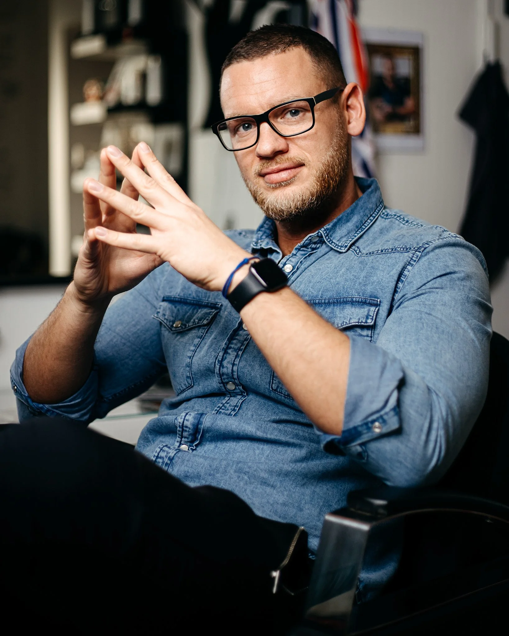 A man with glasses and a beard, wearing a denim shirt, sitting in an office, looking at the camera with his hands clasped together.