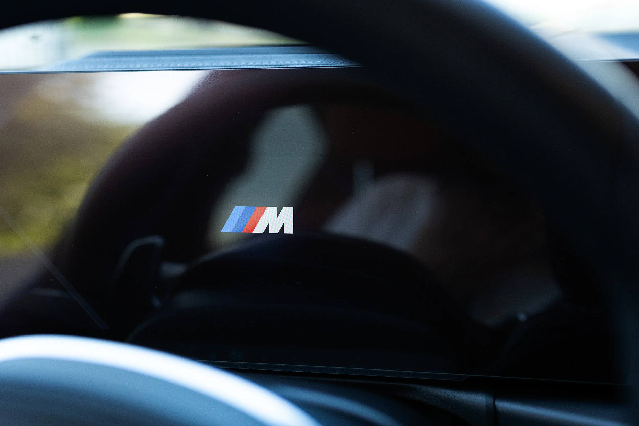 Photo taken from inside a vehicle showing the steering wheel and a dashboard with an 'M' logo in the bottom center of the image.