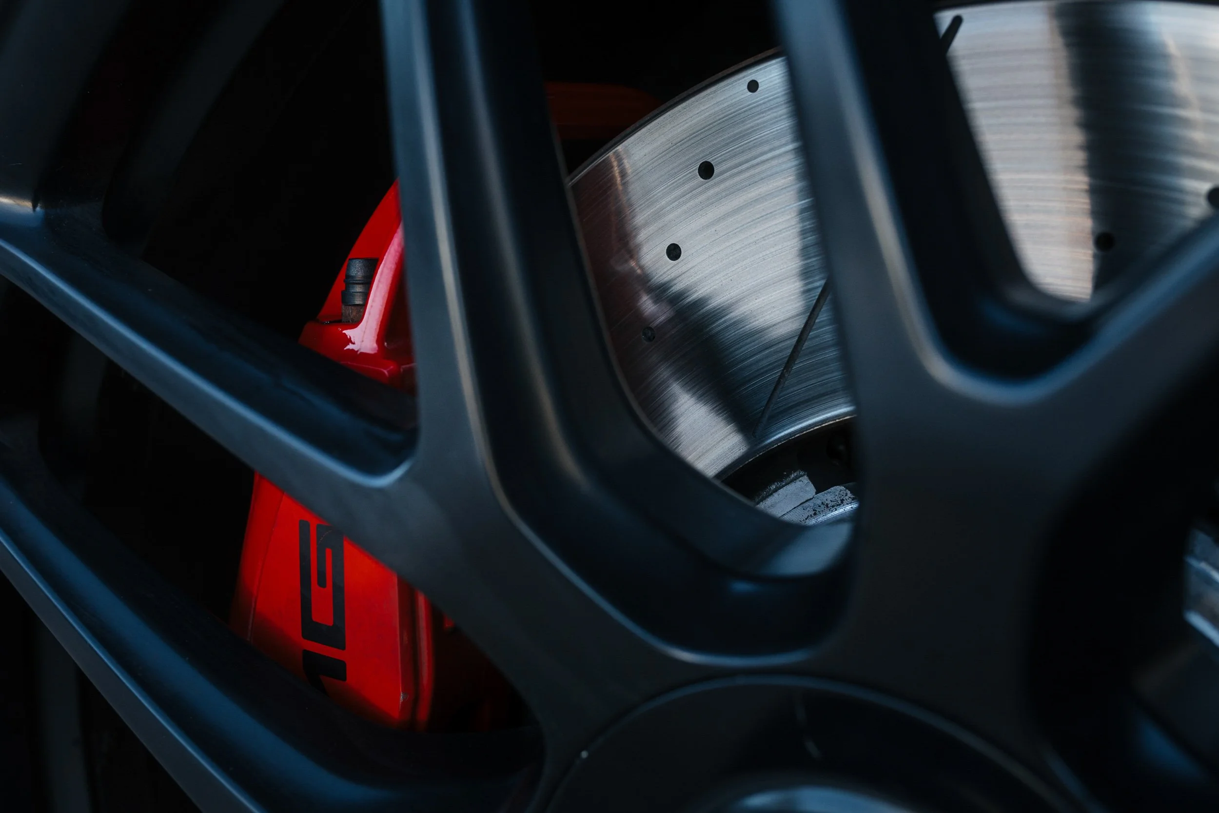 Close-up of a metal disc brake rotor and a red component of a vehicle, seen through black wheel spokes.