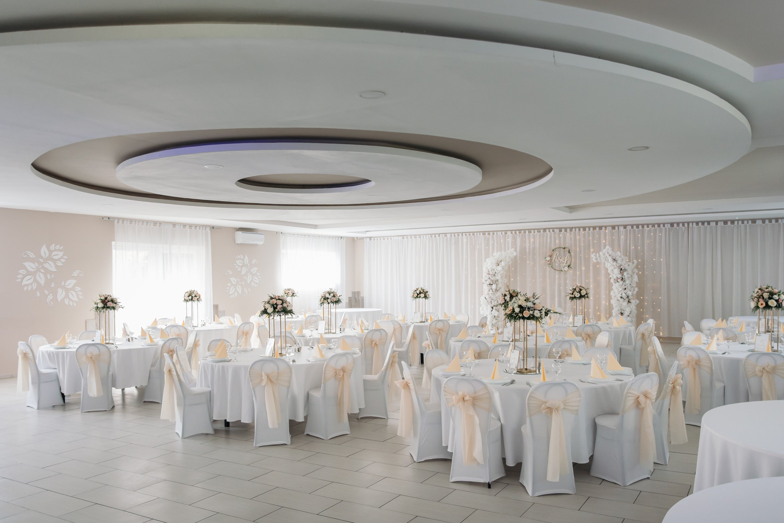 Elegant banquet hall decorated for a wedding reception with round tables covered in white tablecloths, each set with chairs draped in white fabric and tied with bows, floral centerpieces, and ambient lighting featuring sheer curtains and string light