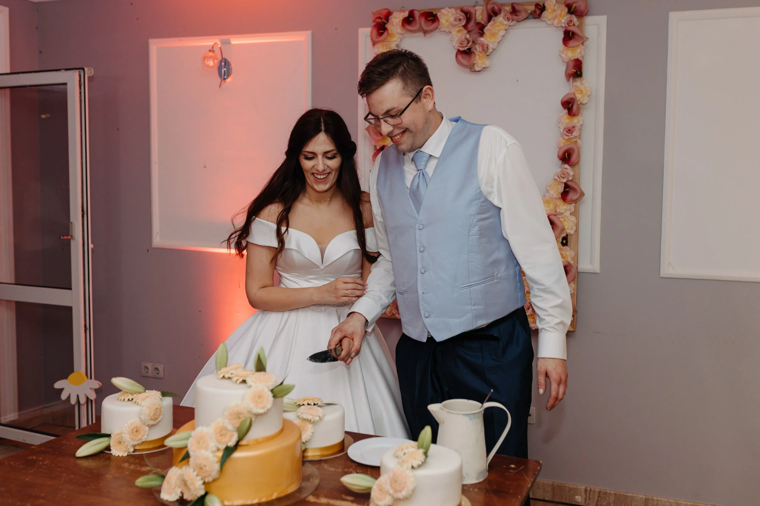 A bride and groom cutting a wedding cake decorated with white and peach flowers, in front of a floral heart decoration on the wall.