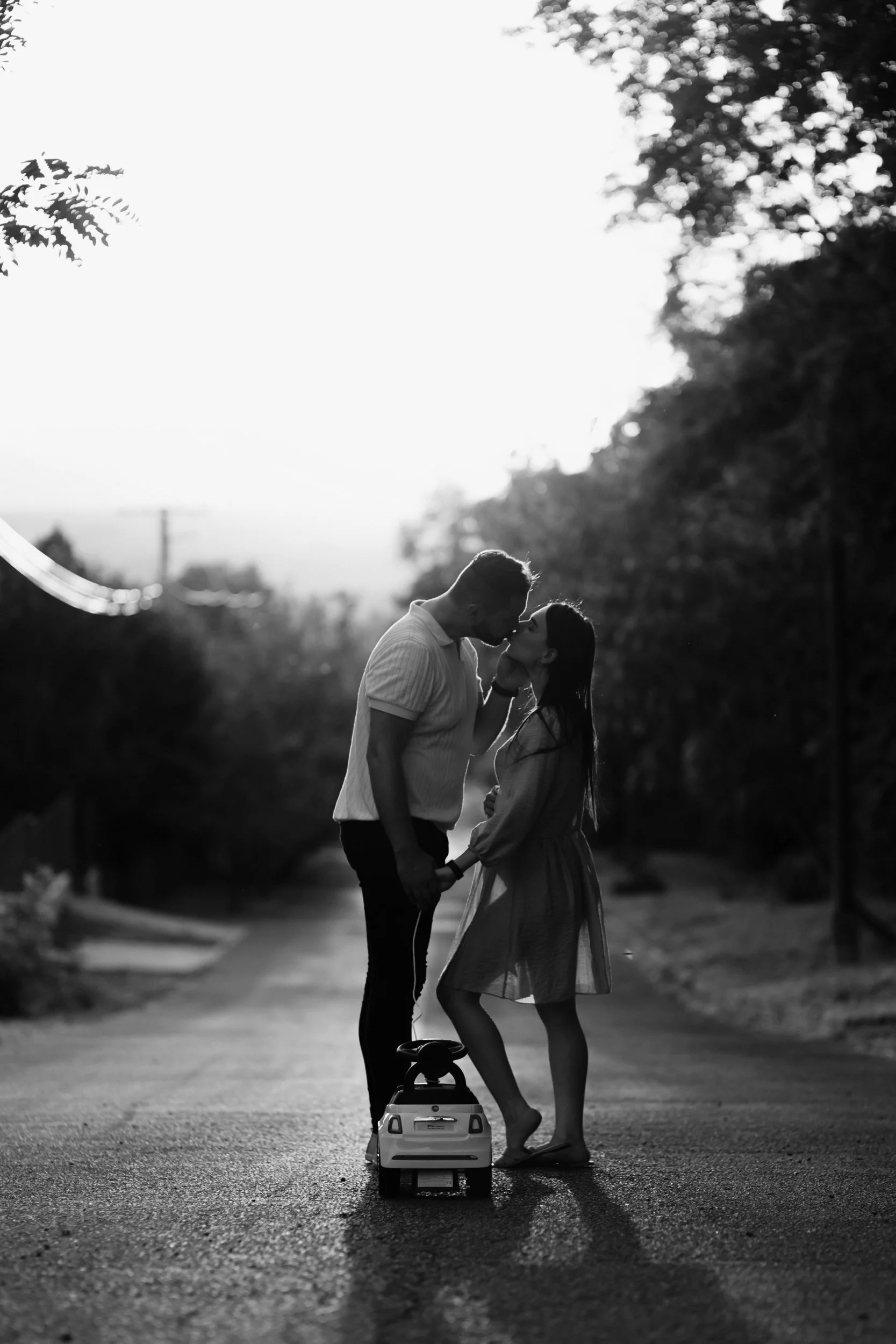 A black and white photo of a couple standing close together on a street, holding hands and leaning in for a kiss, with one of them on a skateboard. The background is blurred with trees and sky, creating a romantic atmosphere.