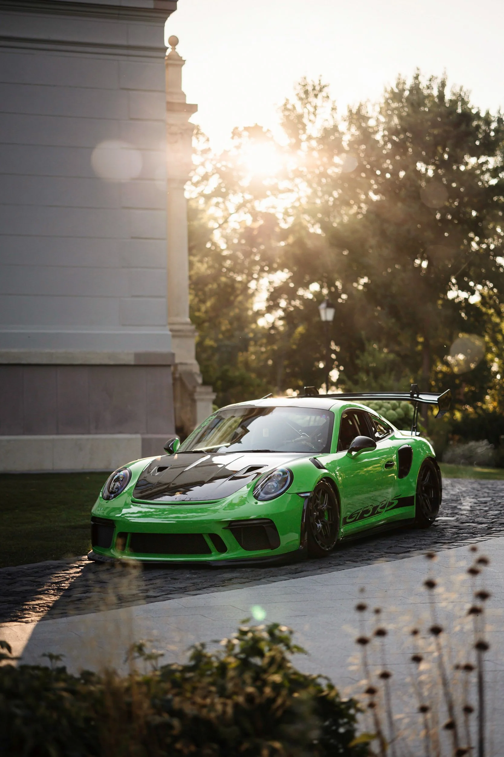 A bright green sports car with black accents and racing decals parked on a cobblestone street during sunset, with a building and trees in the background.