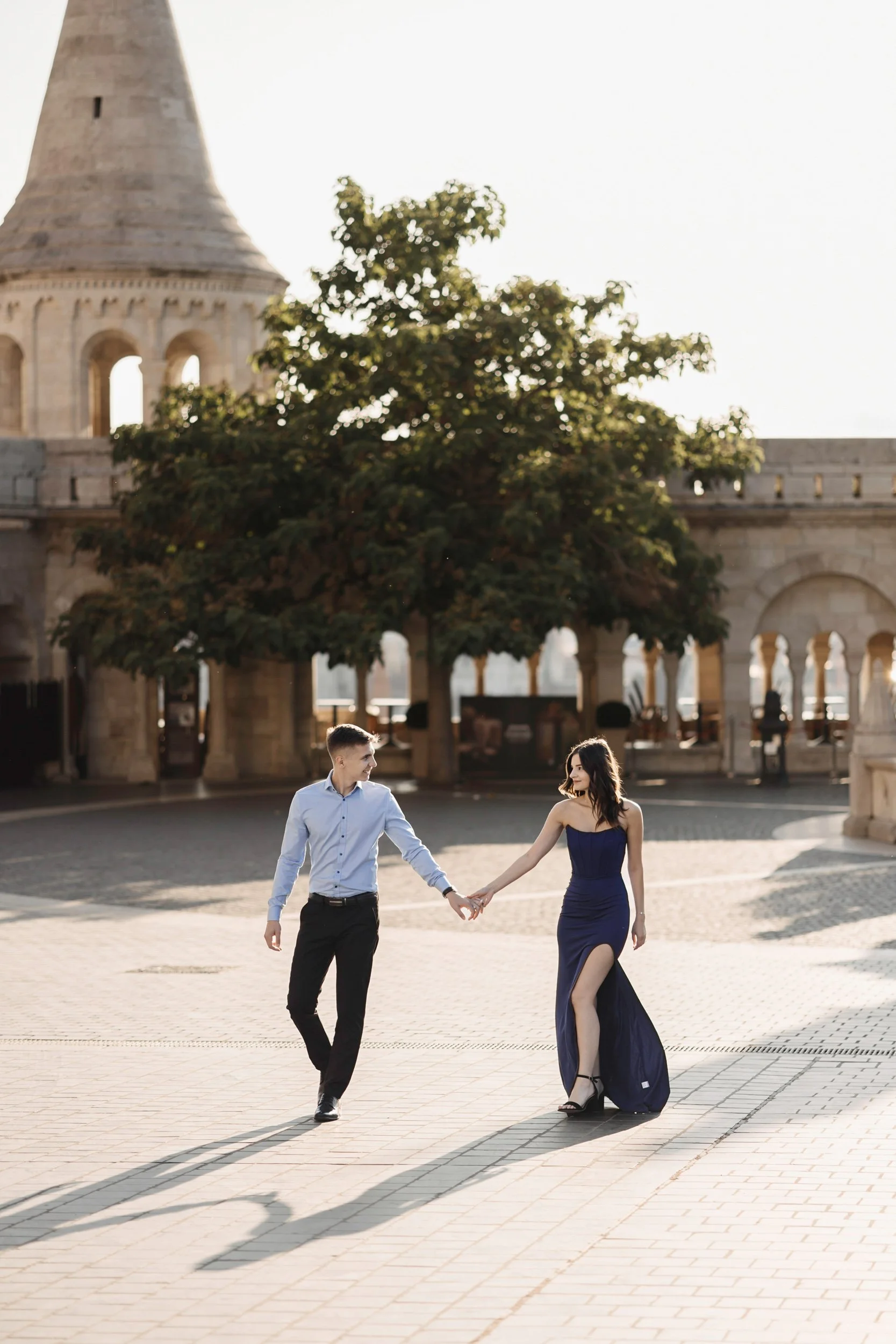 A man and woman holding hands and walking in an outdoor plaza, with a historic stone building and tower in the background during late afternoon sunlight.