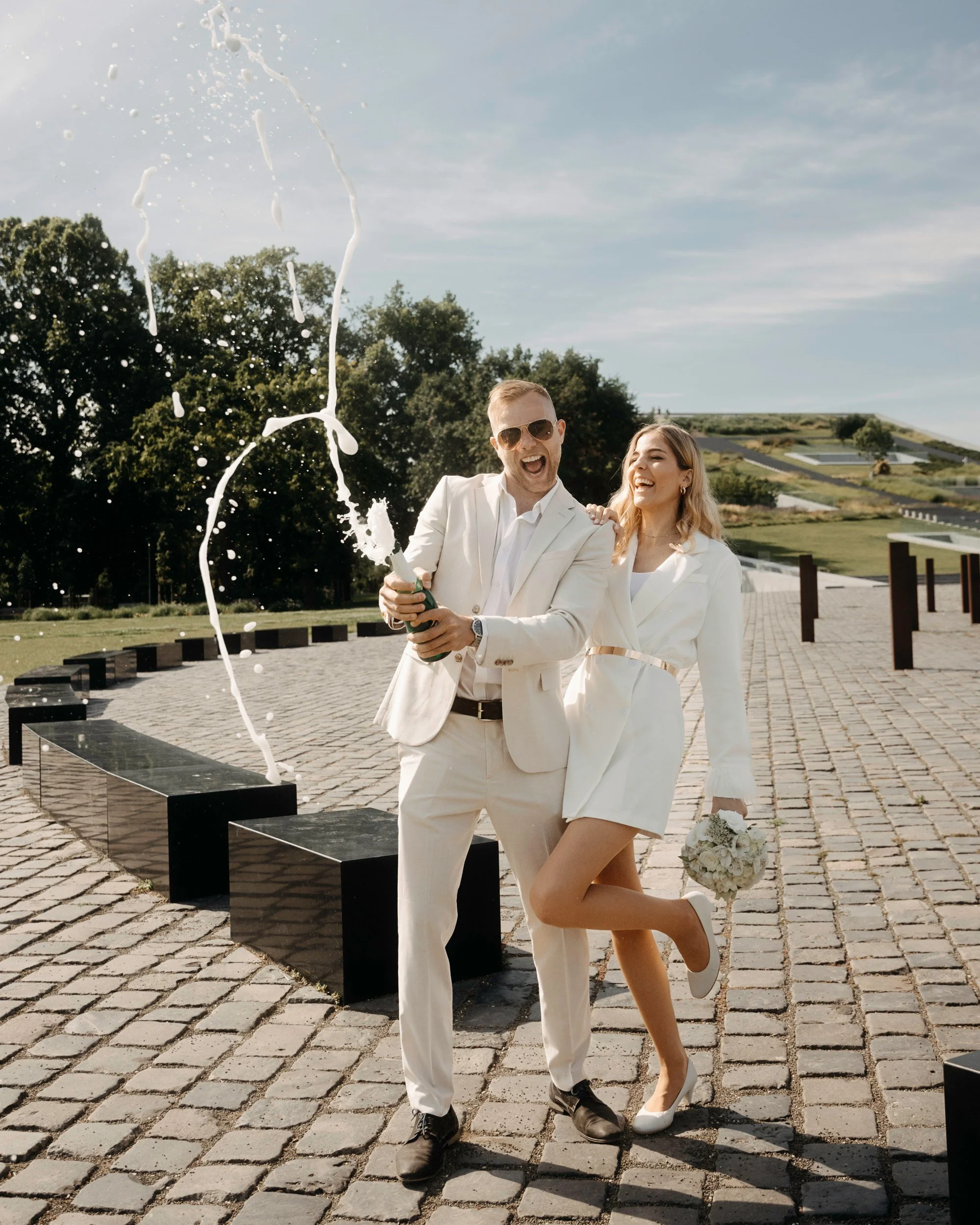 A couple in wedding attire celebrating outdoors on a sunny day, with the man holding a champagne bottle and the woman holding a bouquet of white flowers.