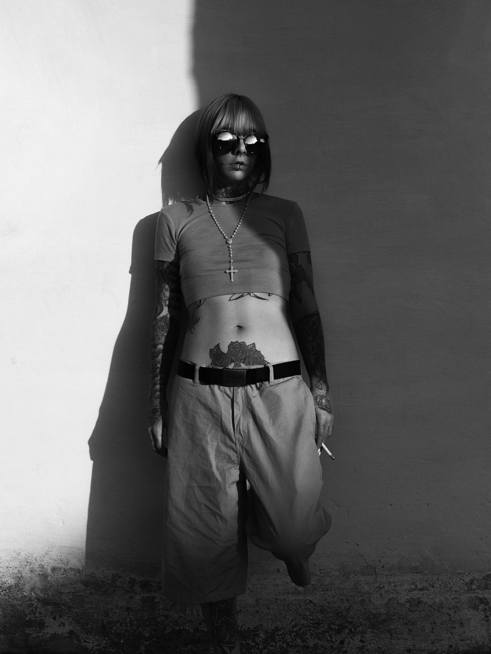 A woman with tattoos on her arms and torso wears sunglasses, a necklace with a cross, a cropped t-shirt, baggy pants, and holds a cigarette while standing near a wall with a shadow cast on it in black and white.