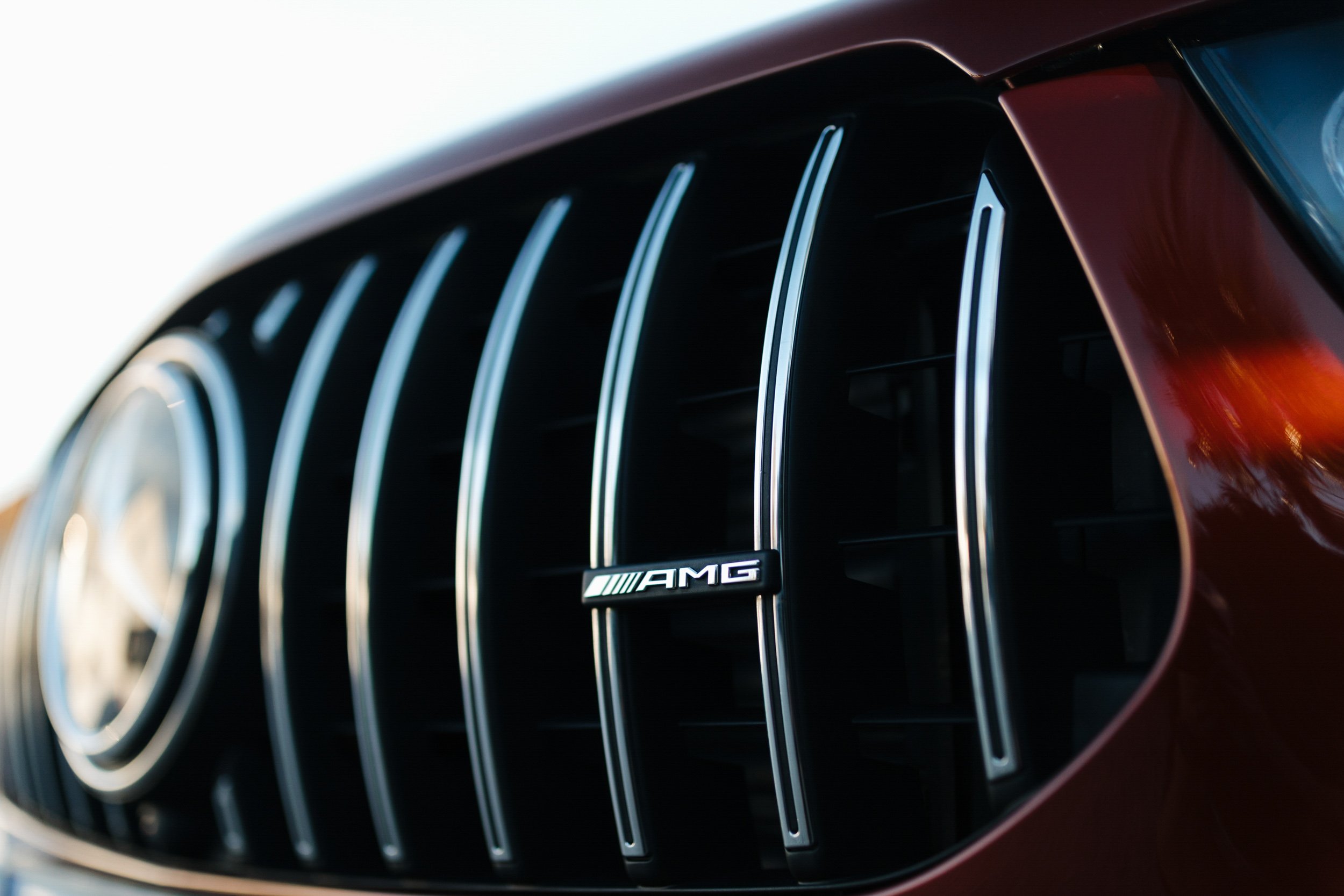 Close-up of a car's front grille with chrome vertical bars and an AMG badge, in a shiny dark red color.