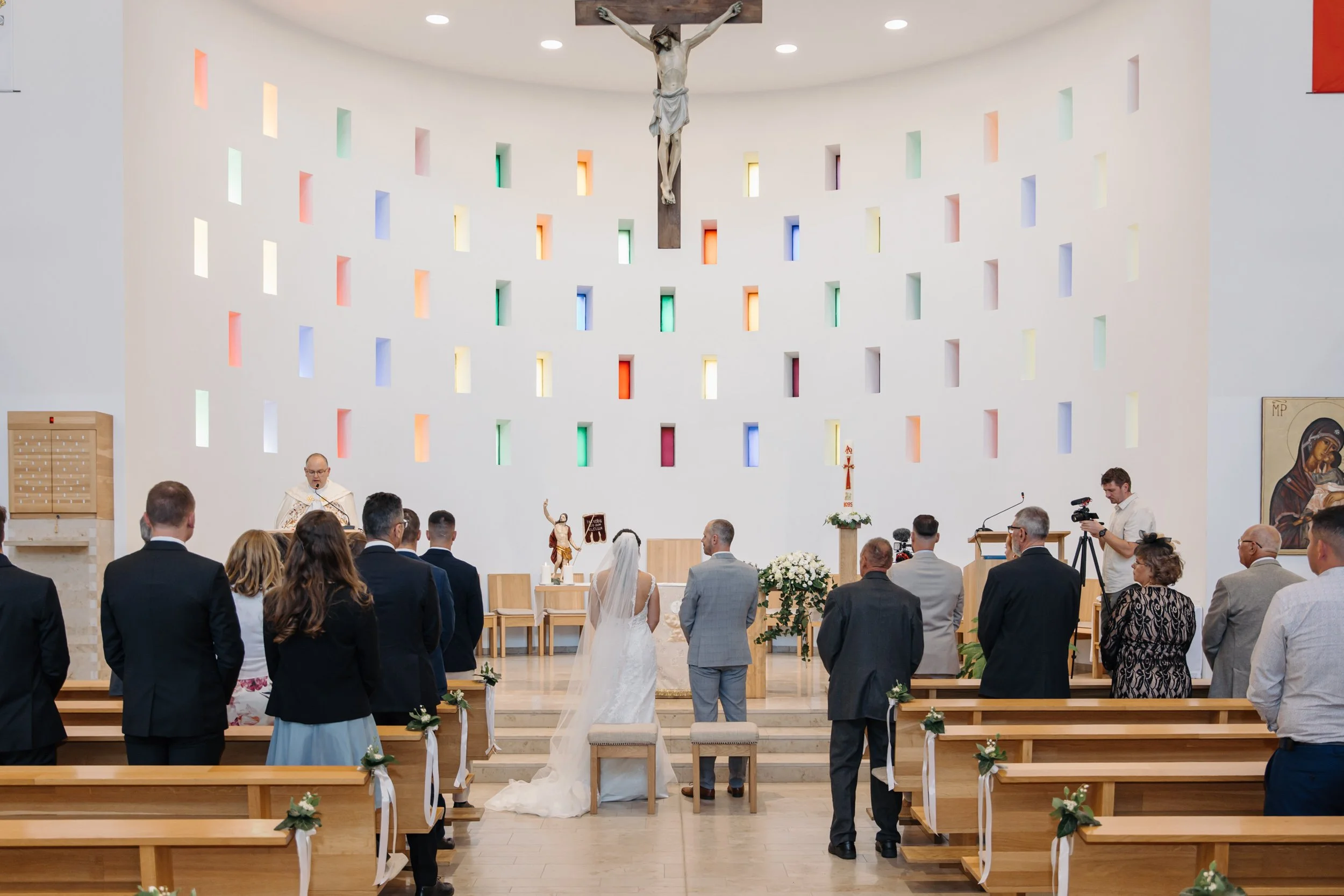 A wedding ceremony inside a church with a white wall, colorful windows, a crucifix, and a group of wedding guests and the bride and groom at the altar.
