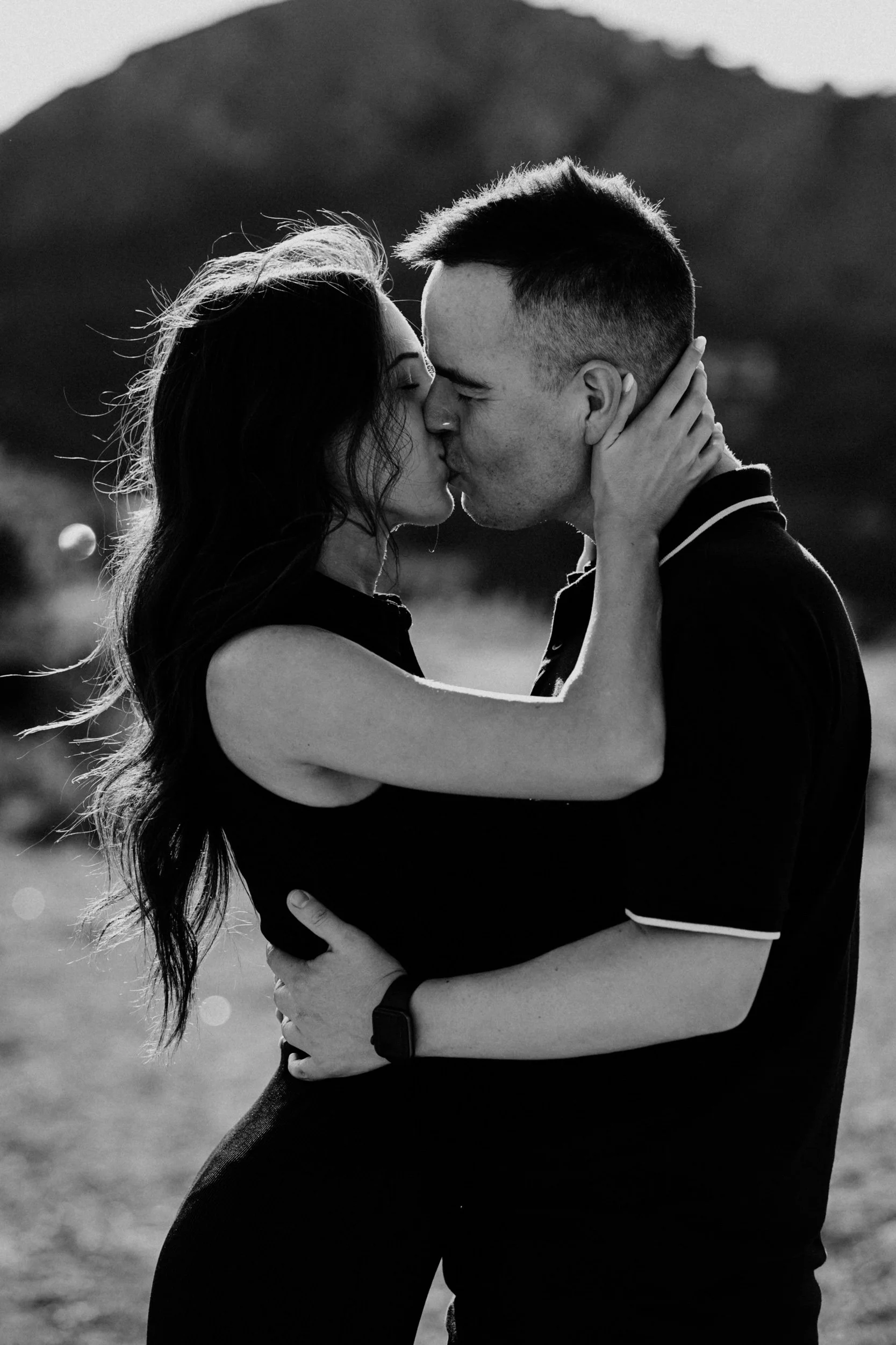 A black and white photo of a couple kissing outdoors, with the woman holding the man's face and the man holding the woman around her waist, mountains blurred in the background.