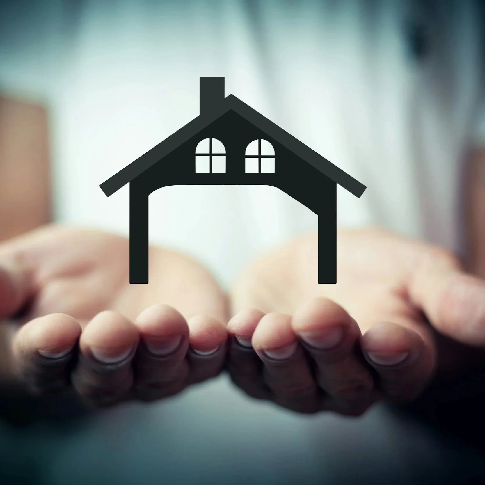 A person's hand holding a small cutout of a house icon with a roof, windows, and chimney, against a blurred background.