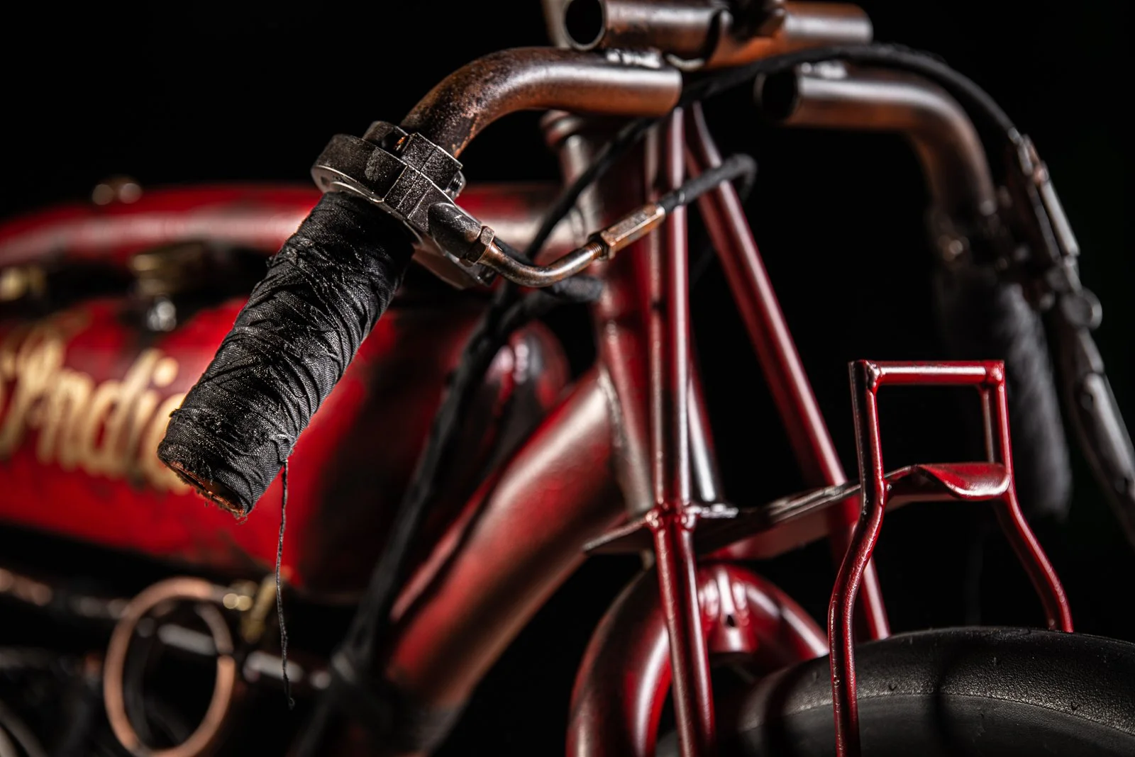 Close-up of a vintage red bicycle with a worn black handlebar grip and a red front rack, with a blurry red and gold sign in the background.
