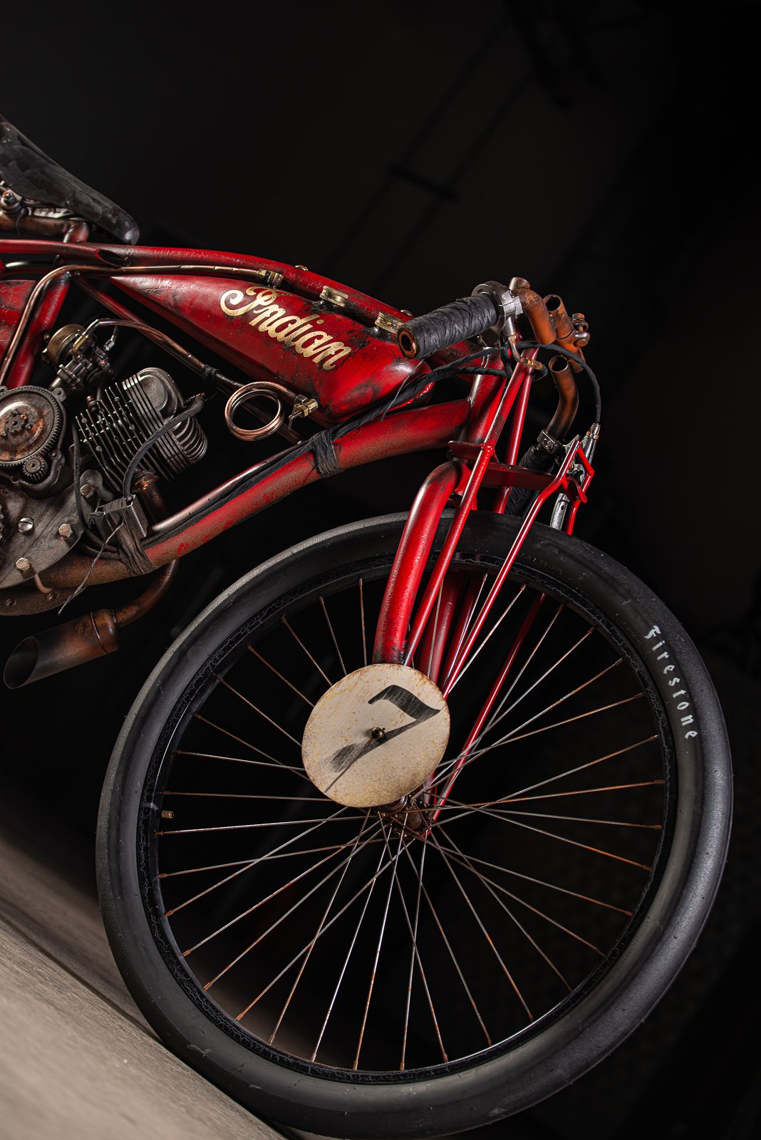Close-up of a vintage red Indian motorcycle with a large front wheel, black handlebar grips, and rusted parts, against a black background.