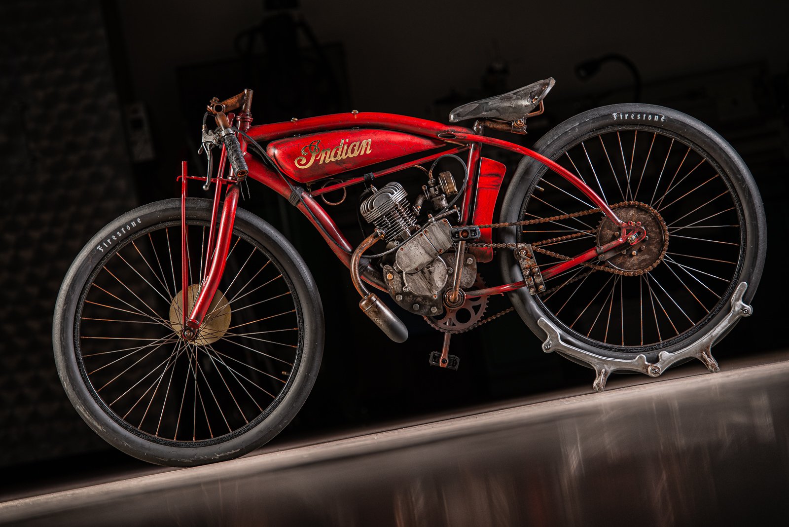A vintage red Indian motorcycle with a visible engine and chain drive, placed on a metal surface against a dark background.