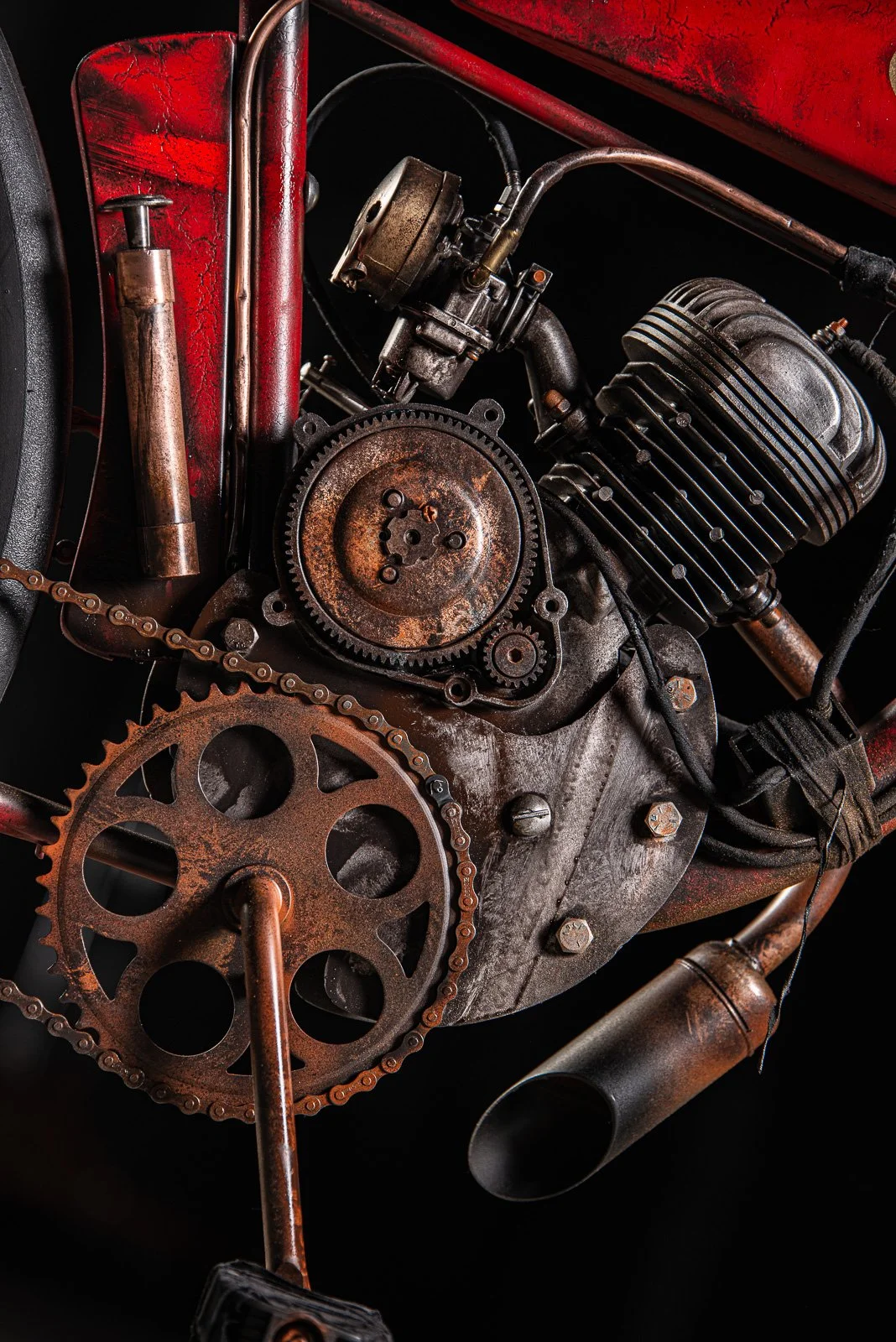 Close-up of a vintage motorcycle engine with rusty gears, chain, and exhaust pipe, against a black background.