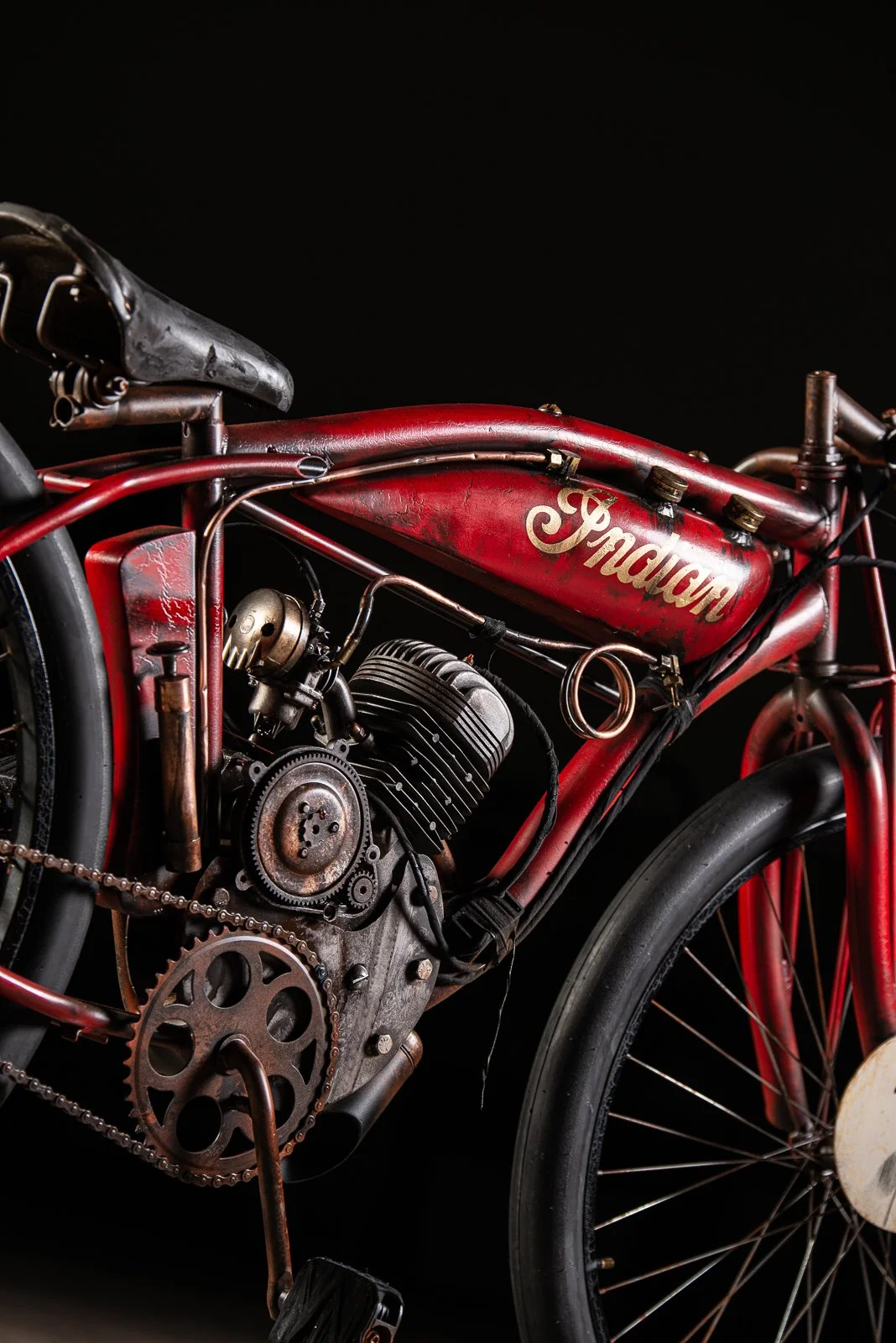 Board track racer red Indian motorcycle against a black background, showing the fuel tank, engine, and front wheel.