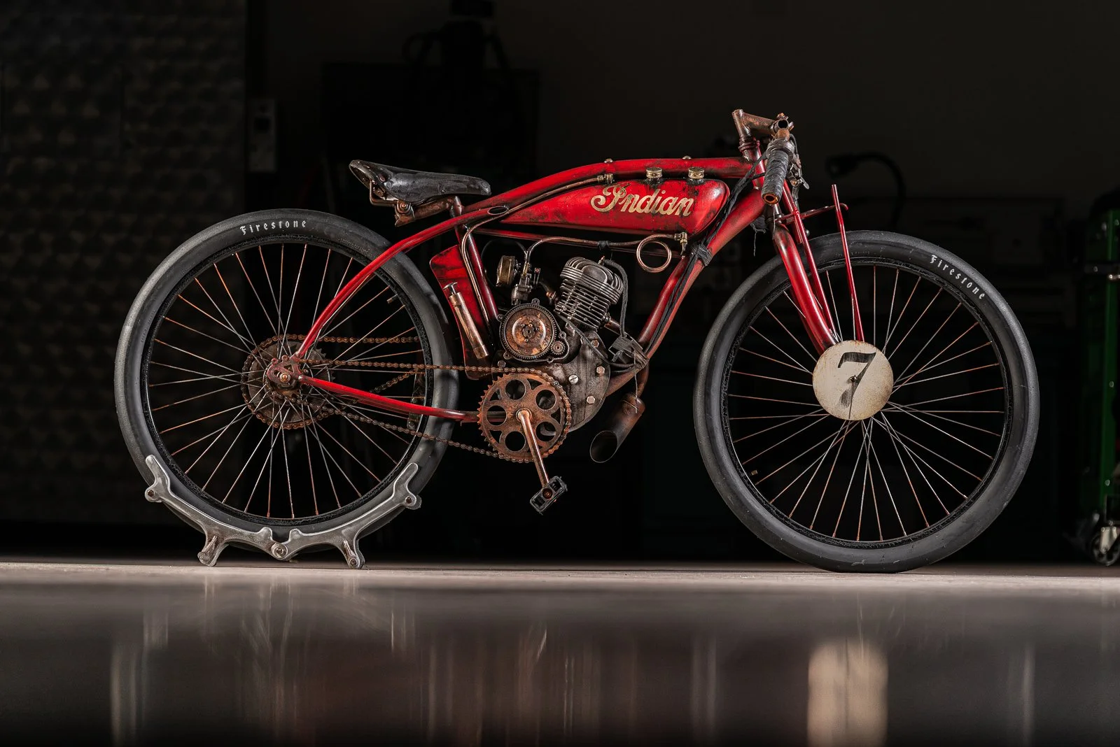 A vintage red motorcycle with the word 'Indian' on the tank, displayed indoors with a black background. The motorcycle has large black tires, with the front tire marked with a white circular plate with the number 7. The bike shows signs of wear and r