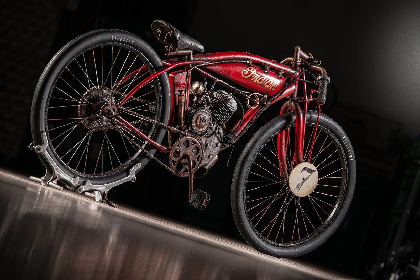 Board track racer red Indian motorcycle with a chain drive, black tires, and a racing number 7 on the front wheel, displayed on a metal stand against a black background.
