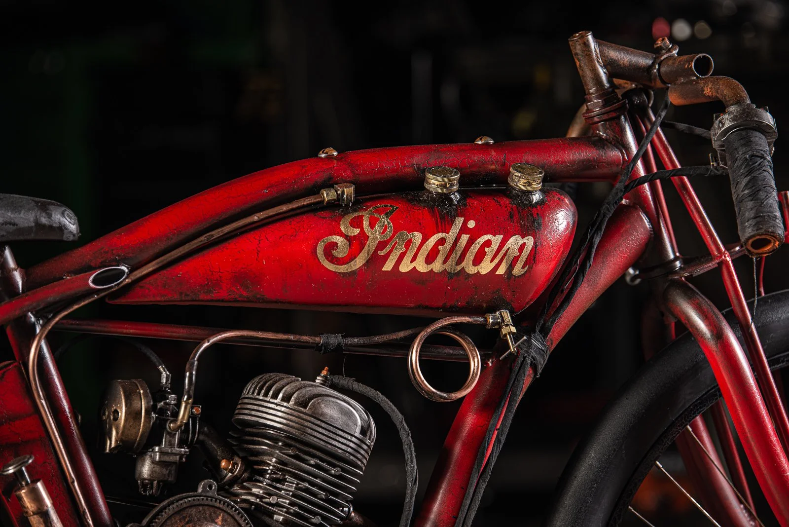 Close-up photo of a vintage red Indian motorcycle with rust and wear, showing the brand logo on the tank, engine, and parts of the frame.