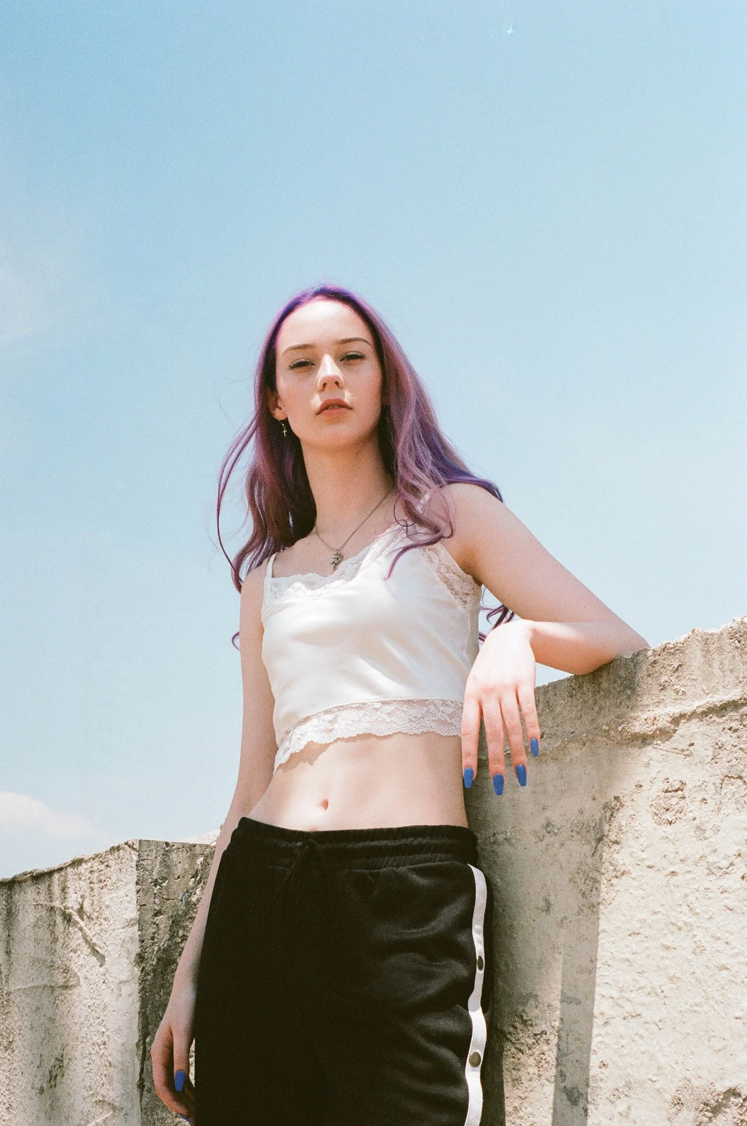 A young woman with purple hair leaning against a concrete wall outdoors with a clear blue sky behind her.